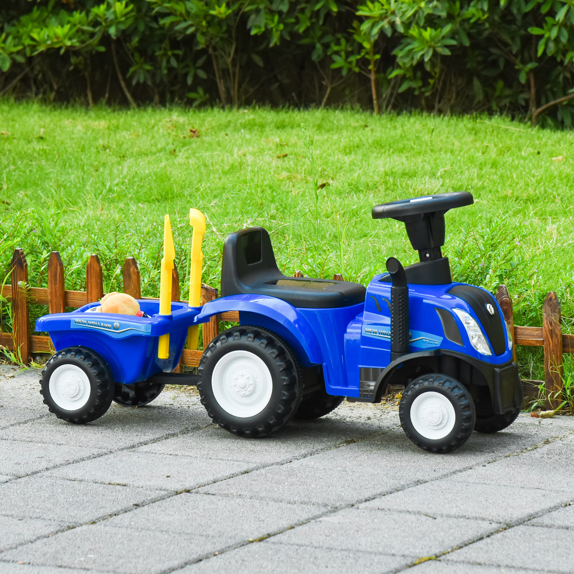 Un trattore giocattolo blu con un rimorchio su una superficie pavimentata, vicino a una staccionata di legno ed erba verde.