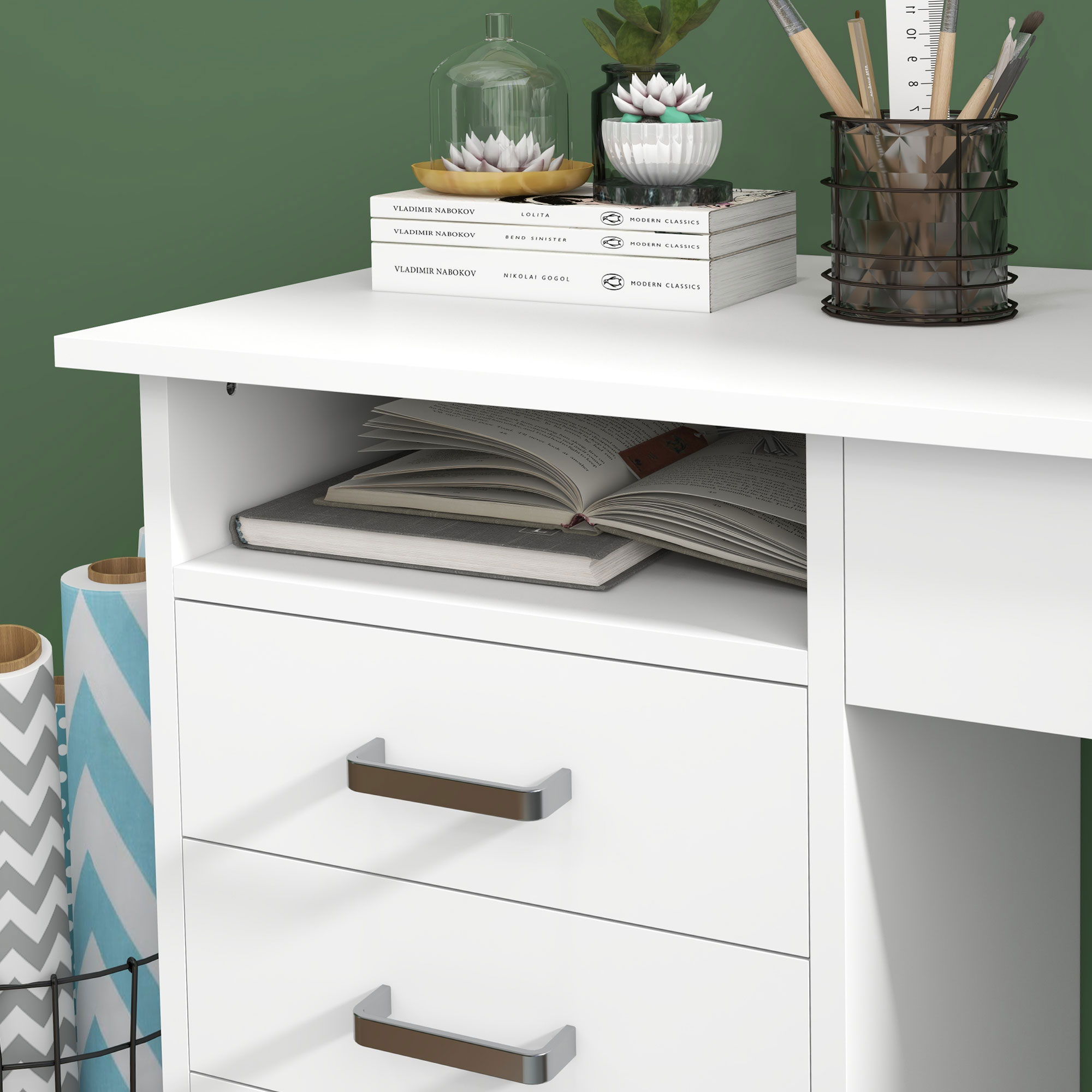 Scrivania bianca con cassetti e mensole, libri, decorazioni e sfondo verde.