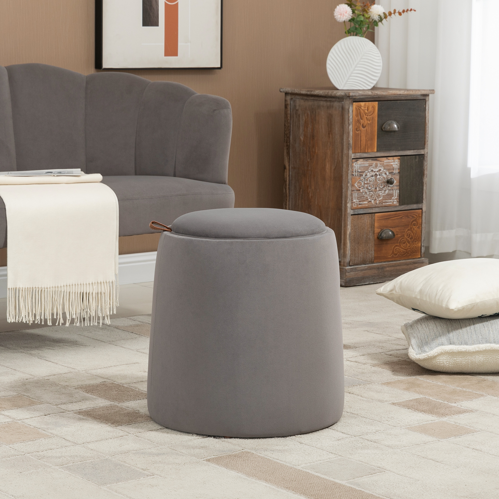 Un pouf in velluto grigio con piano contenitore. Una coperta bianca e mobili in legno sullo sfondo.