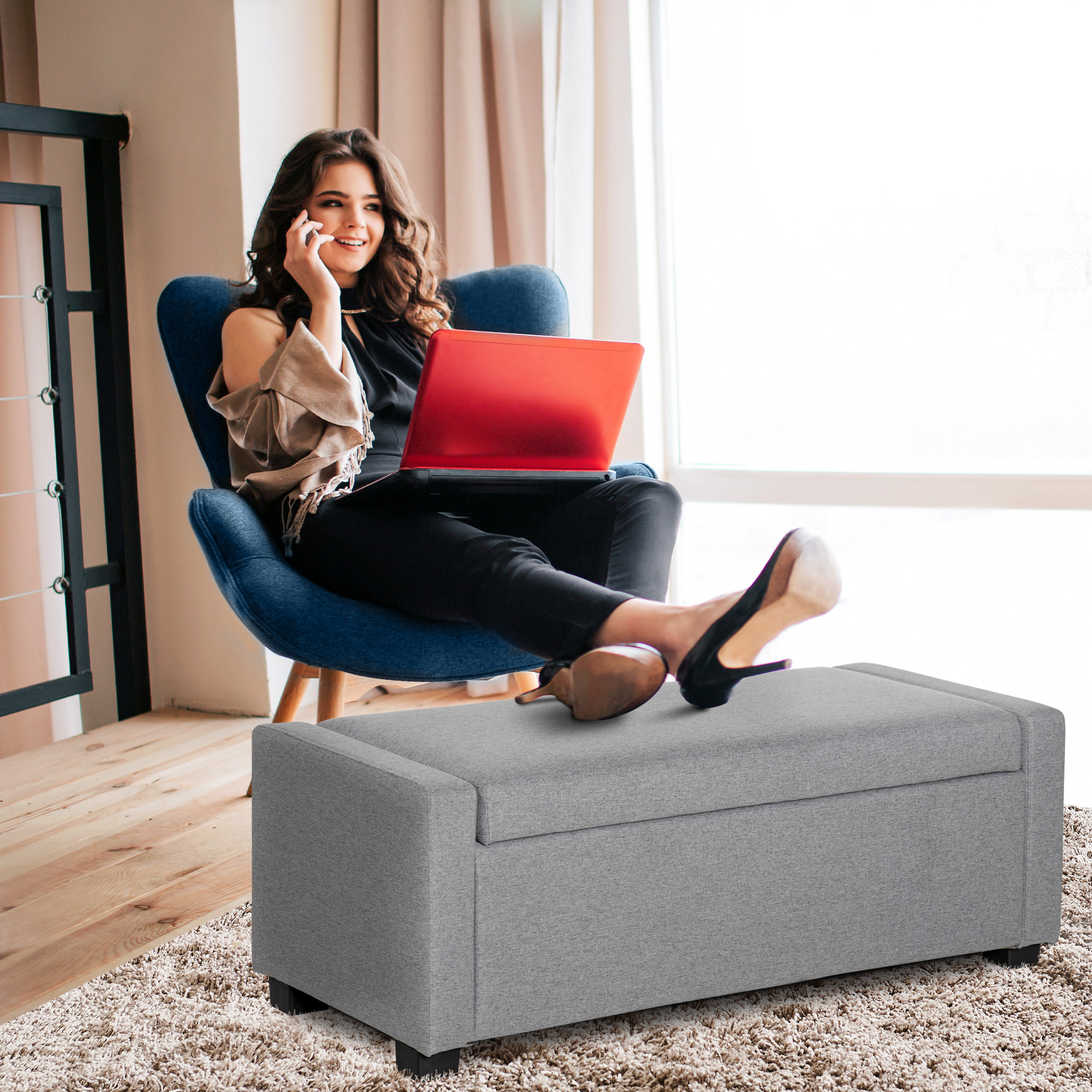 Donna su sedia blu usa laptop, piedi su pouf contenitore grigio.