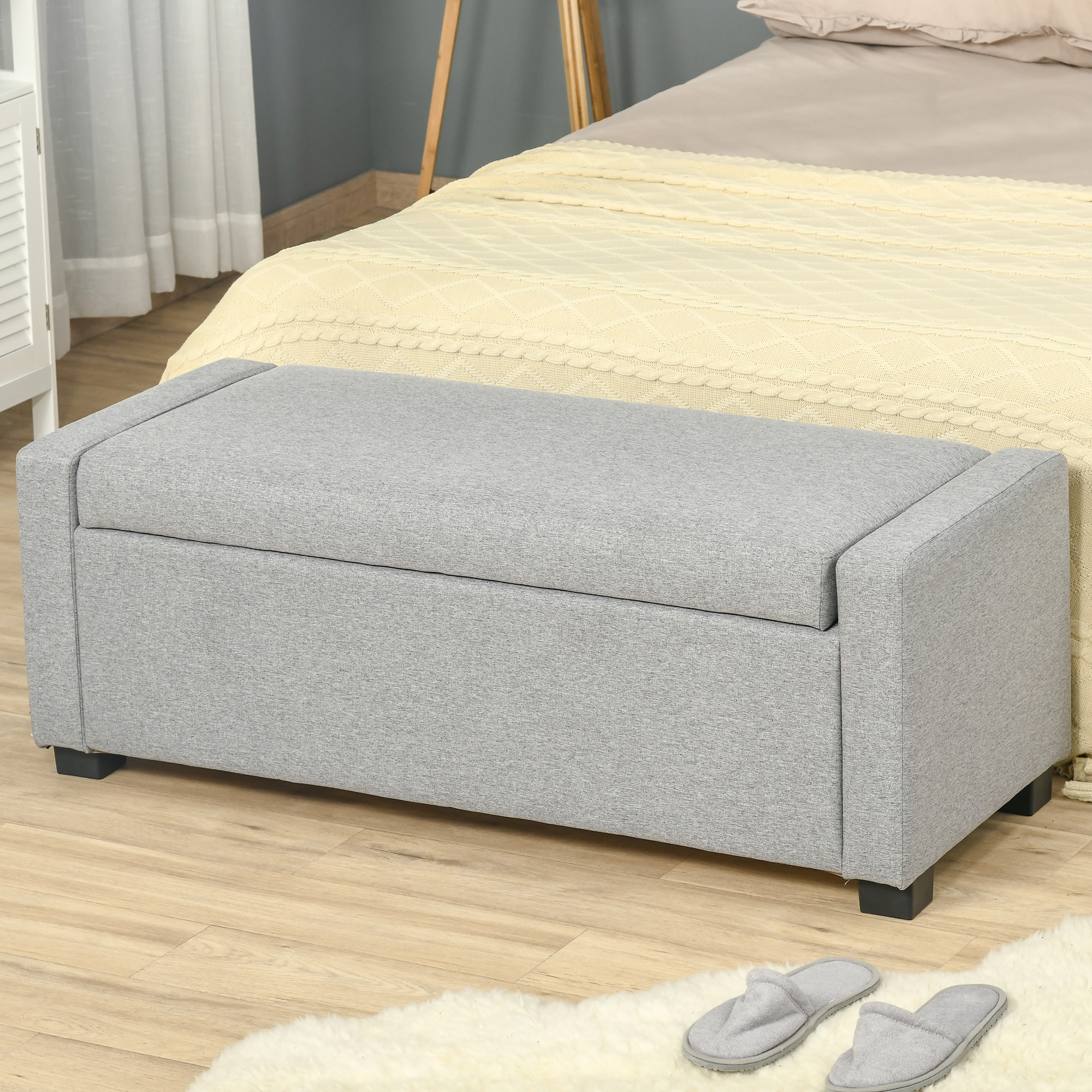 Panca contenitore grigia con letto, tappeto e pantofole sullo sfondo.