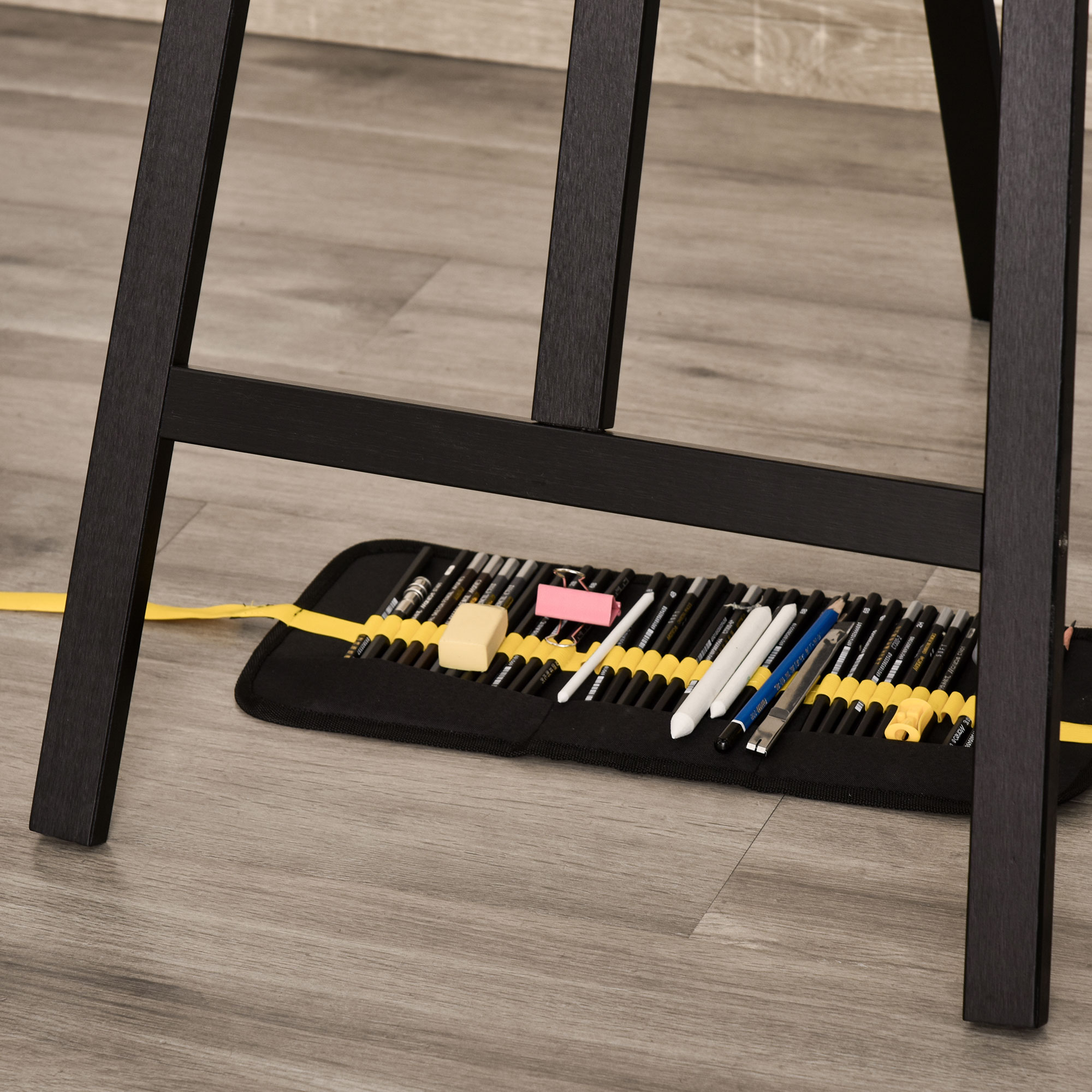 Un cavalletto con strumenti per disegnare, matite e una gomma. Pavimento in legno grigio.