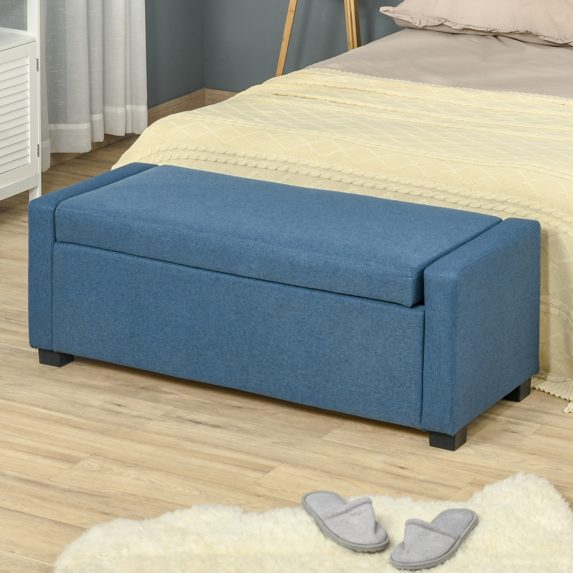 Panca contenitore blu ai piedi di un letto. Le pantofole sono sul pavimento.