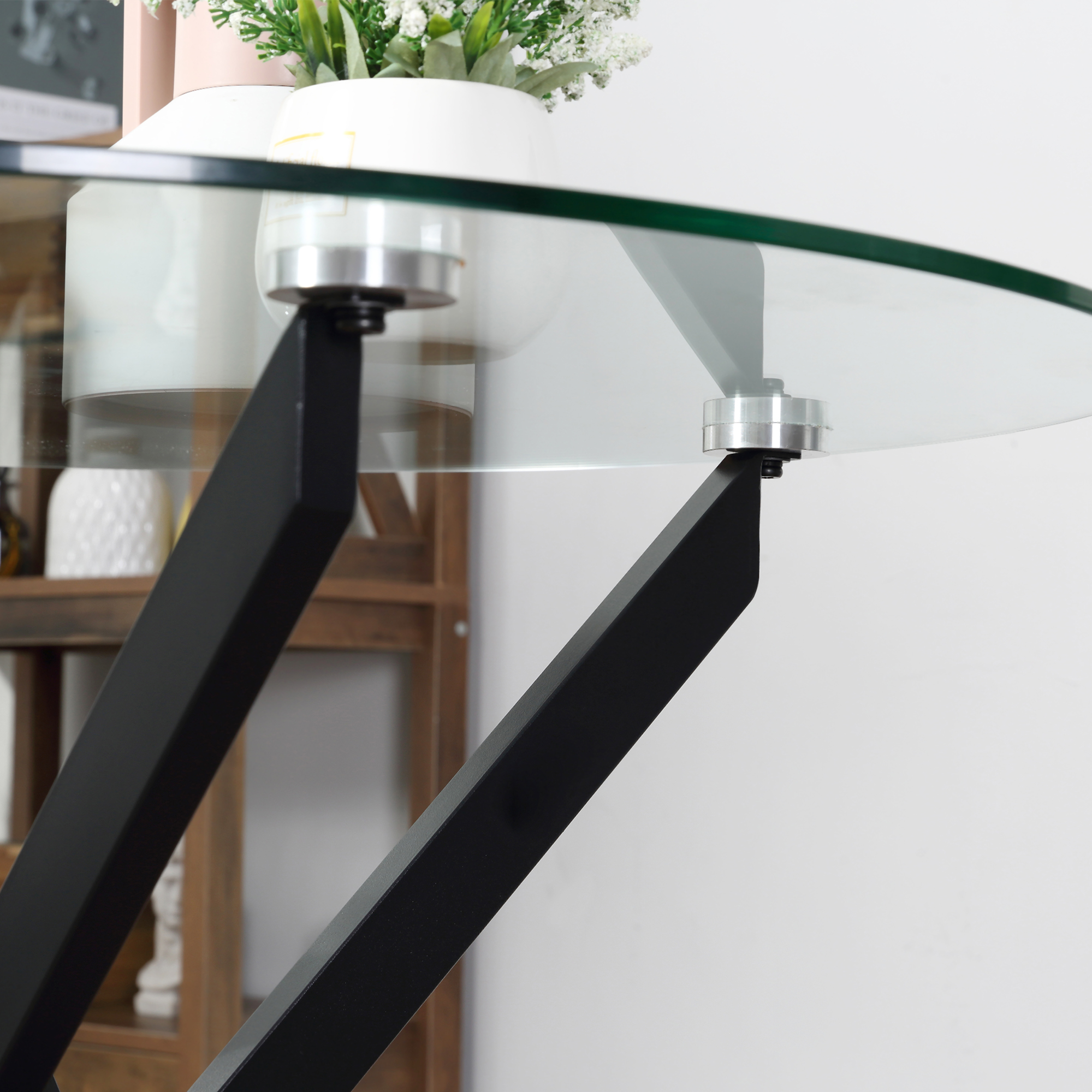 Un tavolo in vetro con gambe in metallo nero, un vaso bianco con piante e una mensola in legno sullo sfondo.
