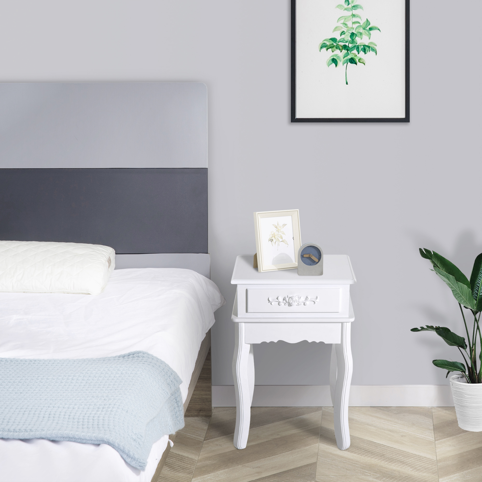 Mesita de noche blanca con cajón y decoración. Cama con cabecero al fondo. Planta, arte y decoración.