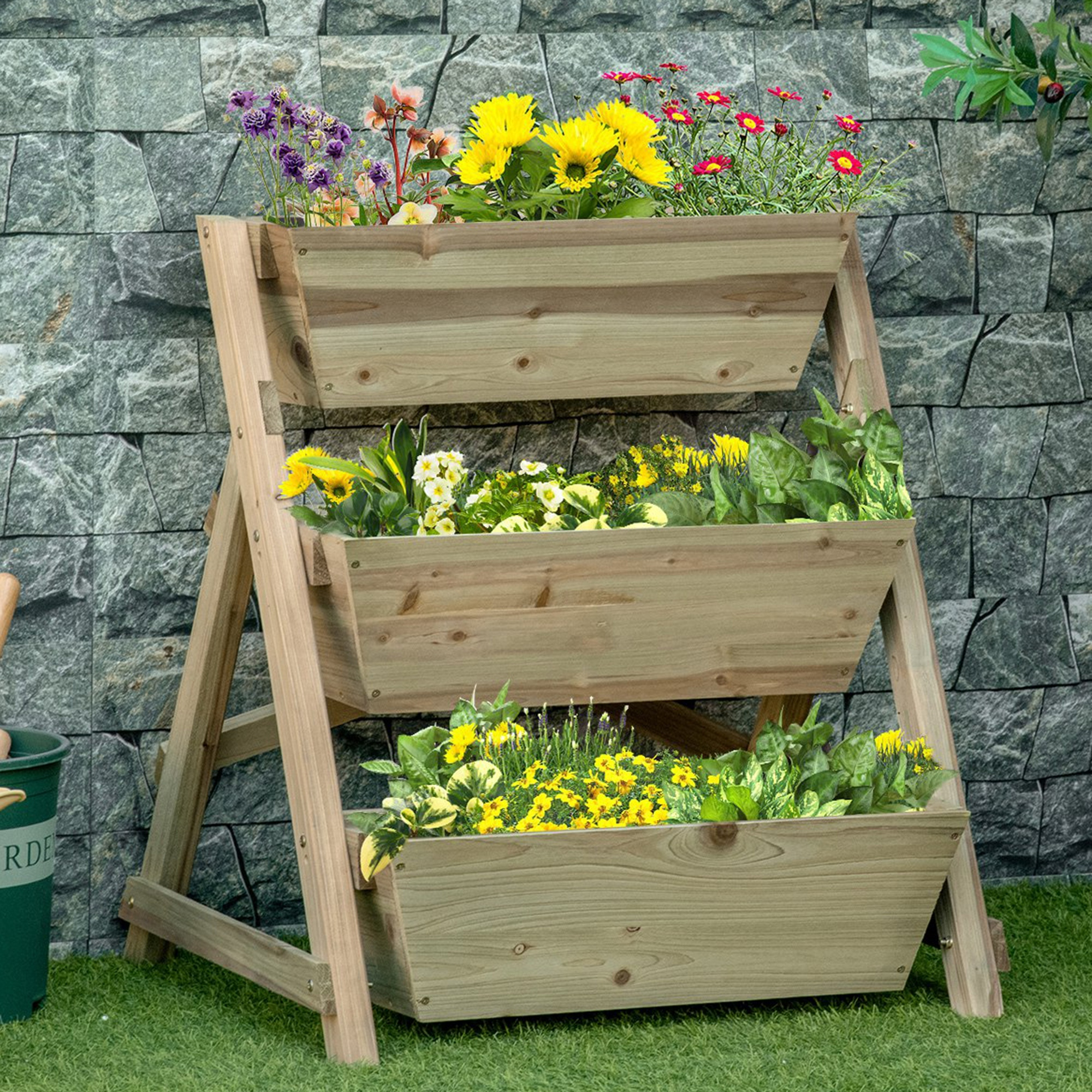 Una jardinera de madera de tres niveles con flores. Se encuentra en césped verde contra muro piedra.