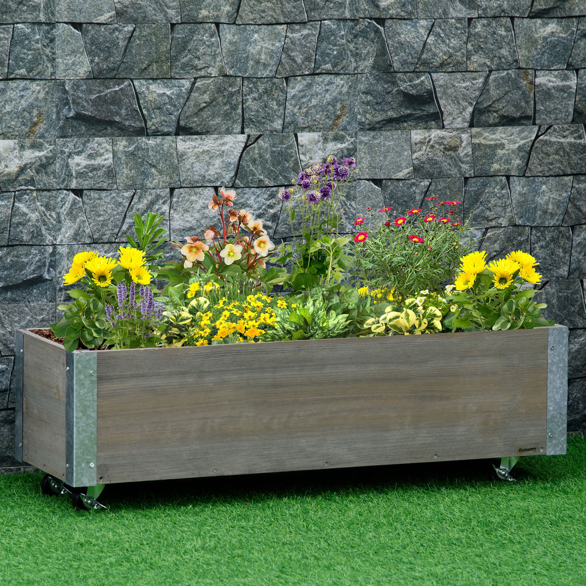Una fioriera in legno grigio con fiori e ruote. Si trova sull'erba verde contro un muro di pietra.