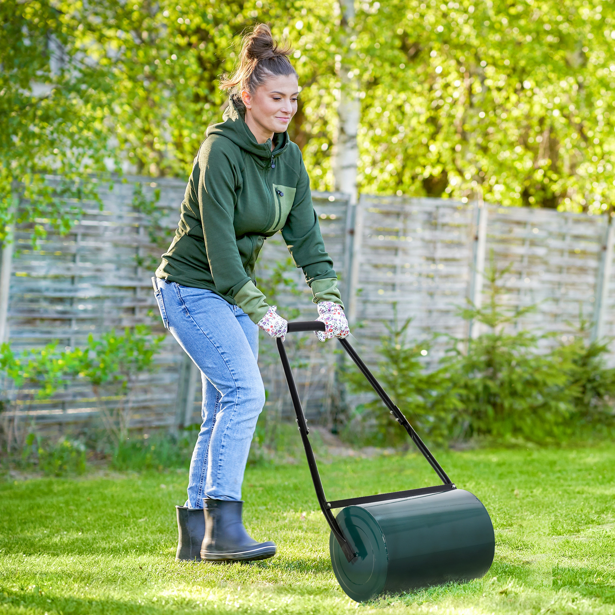 Donna in giacca verde rotola un rullo da prato verde sull'erba. Sfondo: recinzione e alberi verdi.