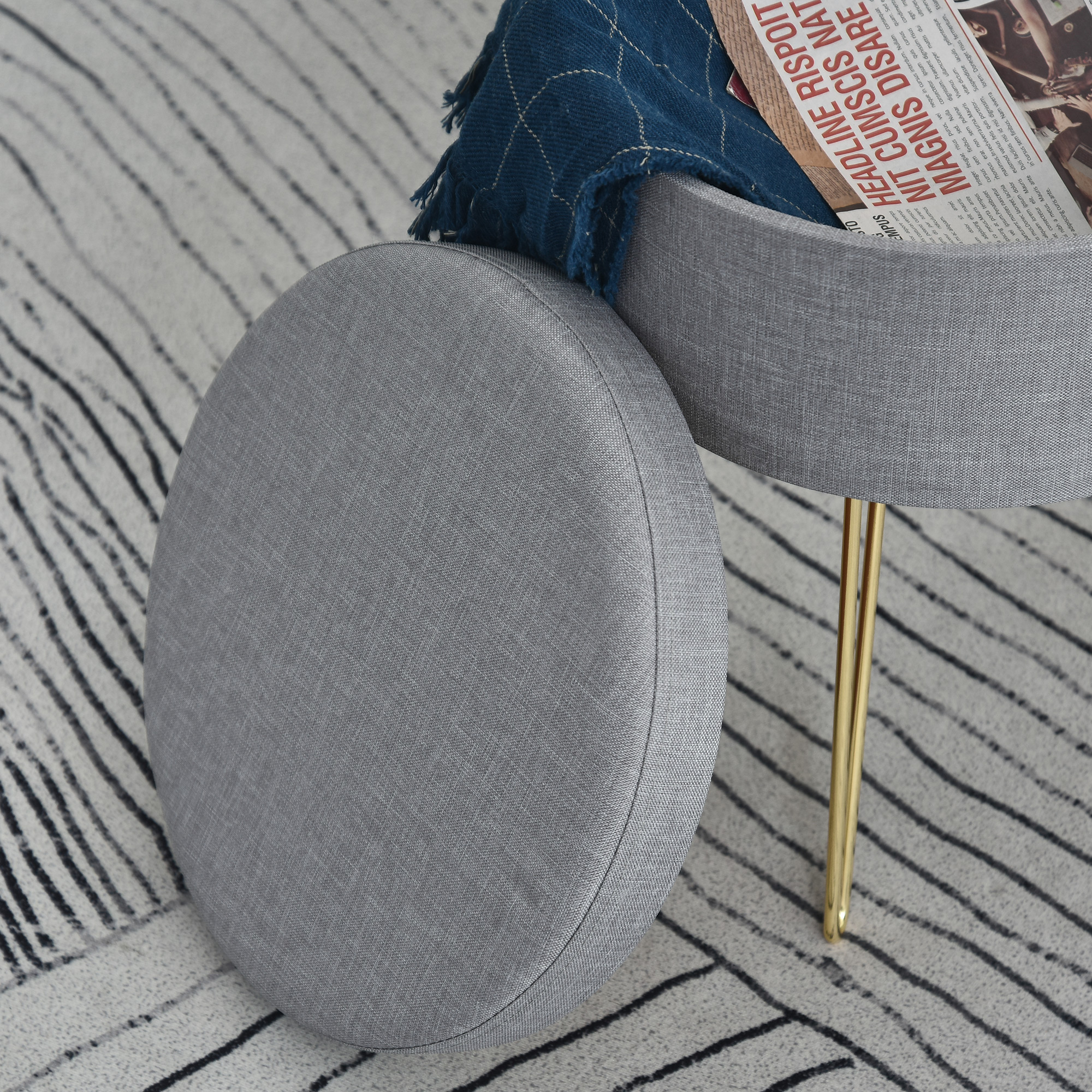 Un pouf rotondo grigio con una coperta blu e un giornale sopra. Gambe dorate visibili. Vista da vicino.