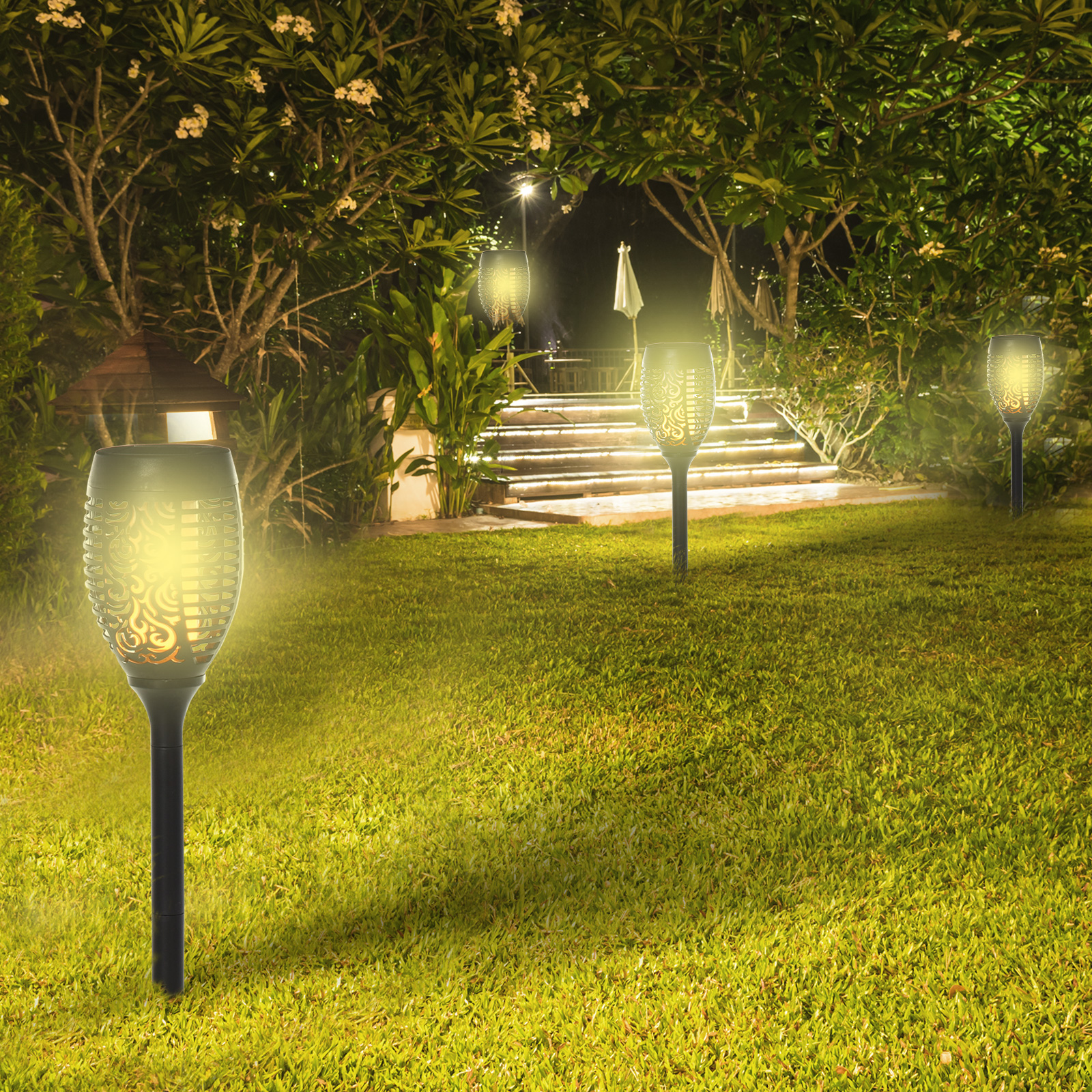 Luci a forma di torcia illuminano un prato erboso di notte. Alberi e una scala sono visibili sullo sfondo.