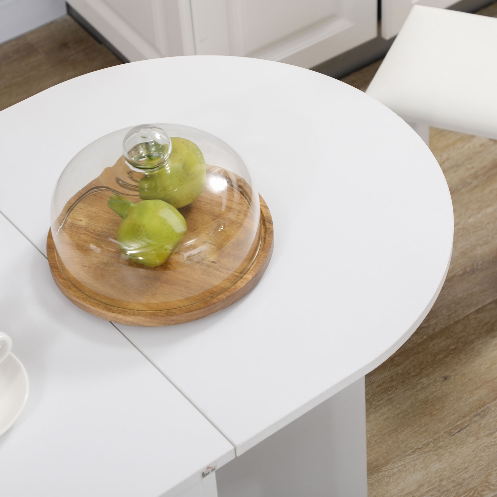 Tavolo bianco con vassoio in legno contenente due pere verdi sotto campana di vetro. Sedia e pavimento bianchi visibili.