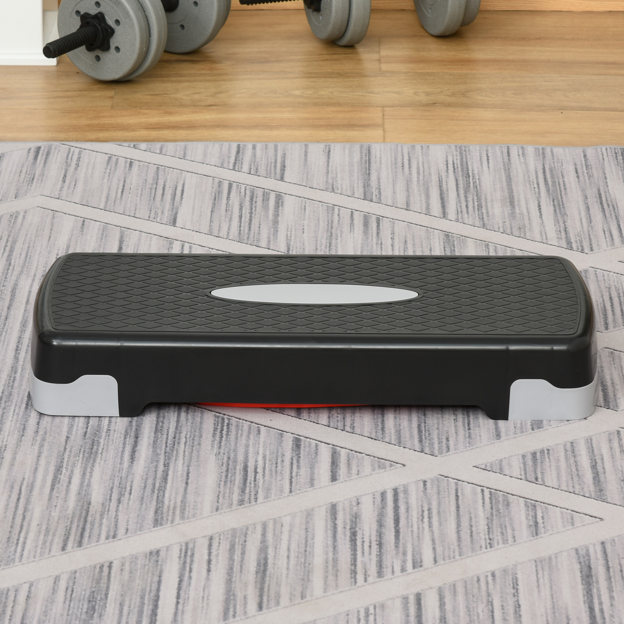 Piattaforma di step nera e grigia su un tappeto. Manubri sullo sfondo. Pavimento in legno.