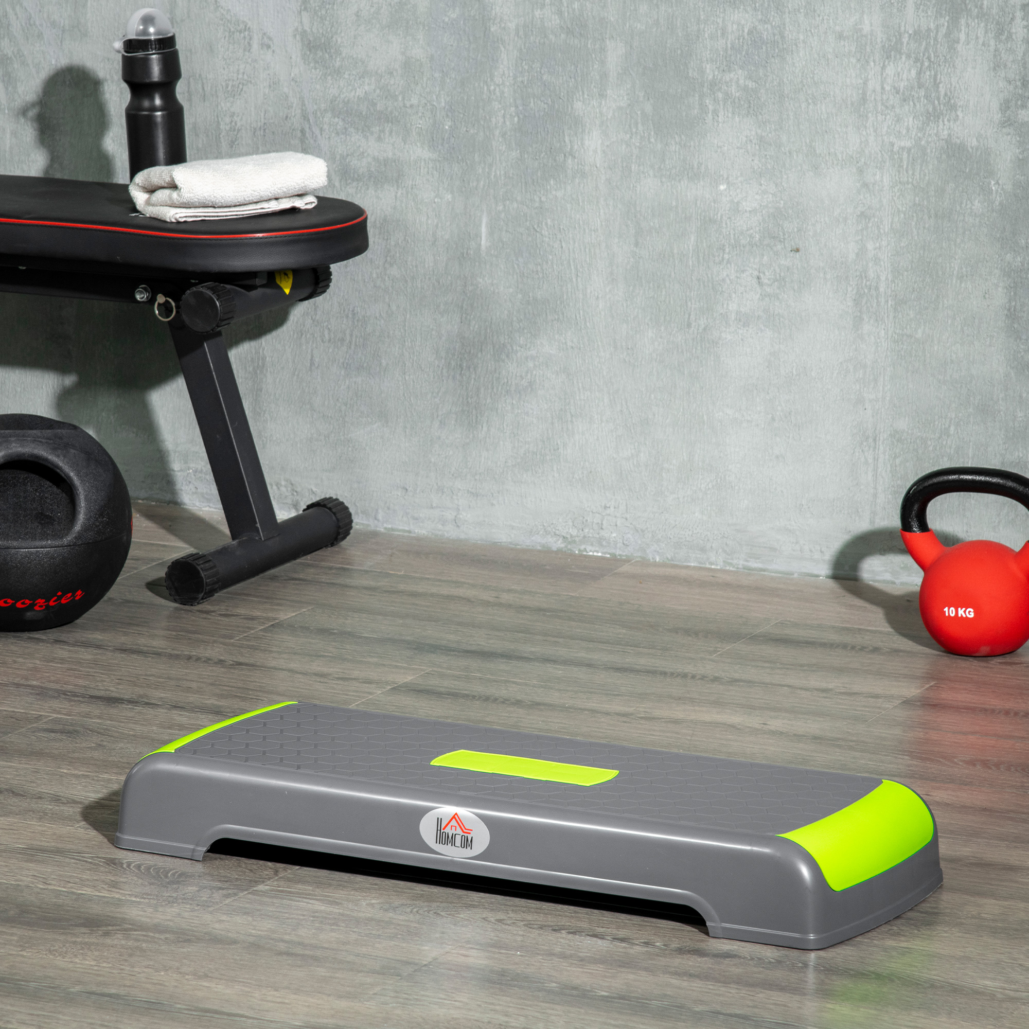 Piattaforma di step grigia e verde su pavimento in legno. Kettlebell, borraccia e panca sullo sfondo.