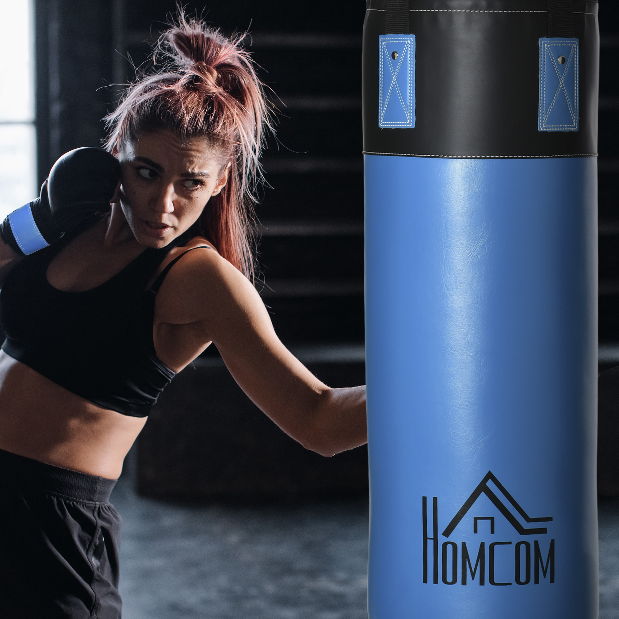 Donna con guantoni da boxe che colpisce un sacco da boxe blu. Ha i capelli rossi.