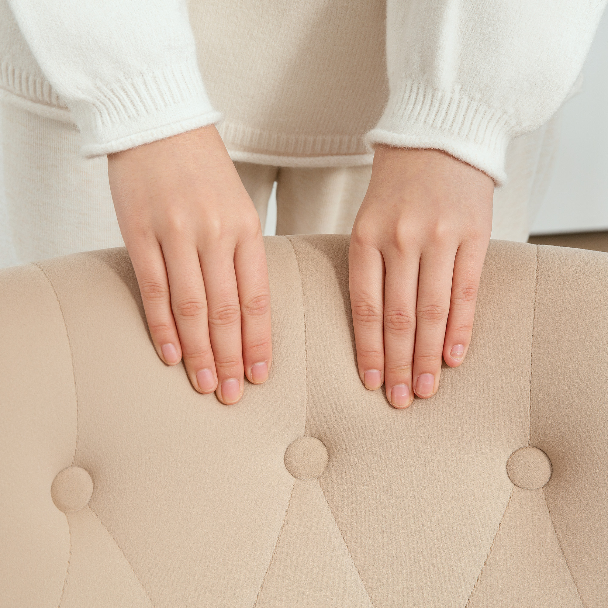 Mani appoggiate su una sedia beige con bottoni. Sfondo chiaro e neutro.