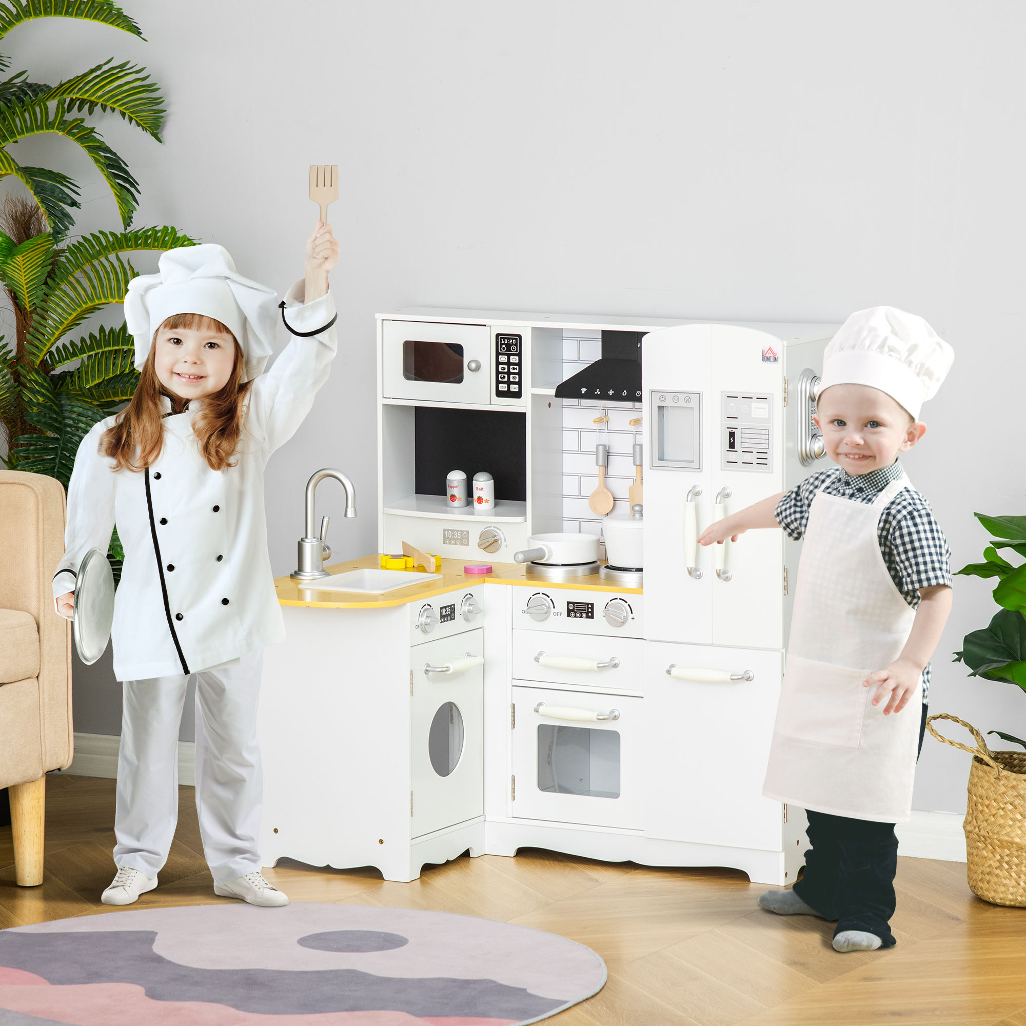 Due bambini vestiti da chef davanti a una cucina giocattolo bianca, con una spatola e un piatto.