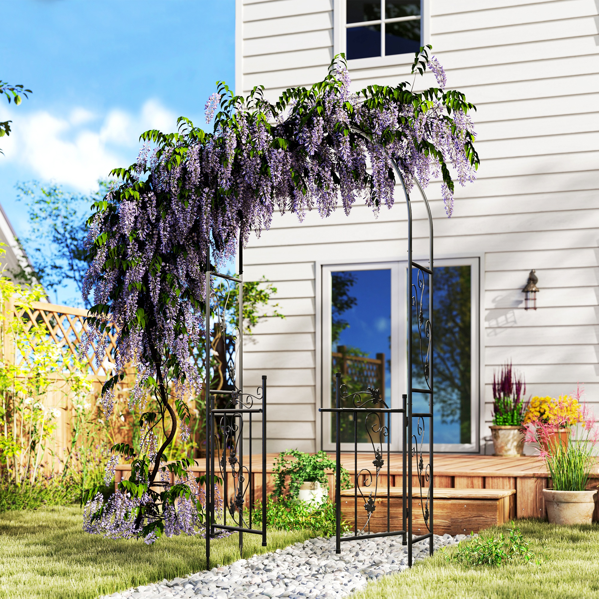 Arco da giardino in metallo nero con fiori viola e un sentiero di pietra. Sfondo casa bianca e cielo blu.