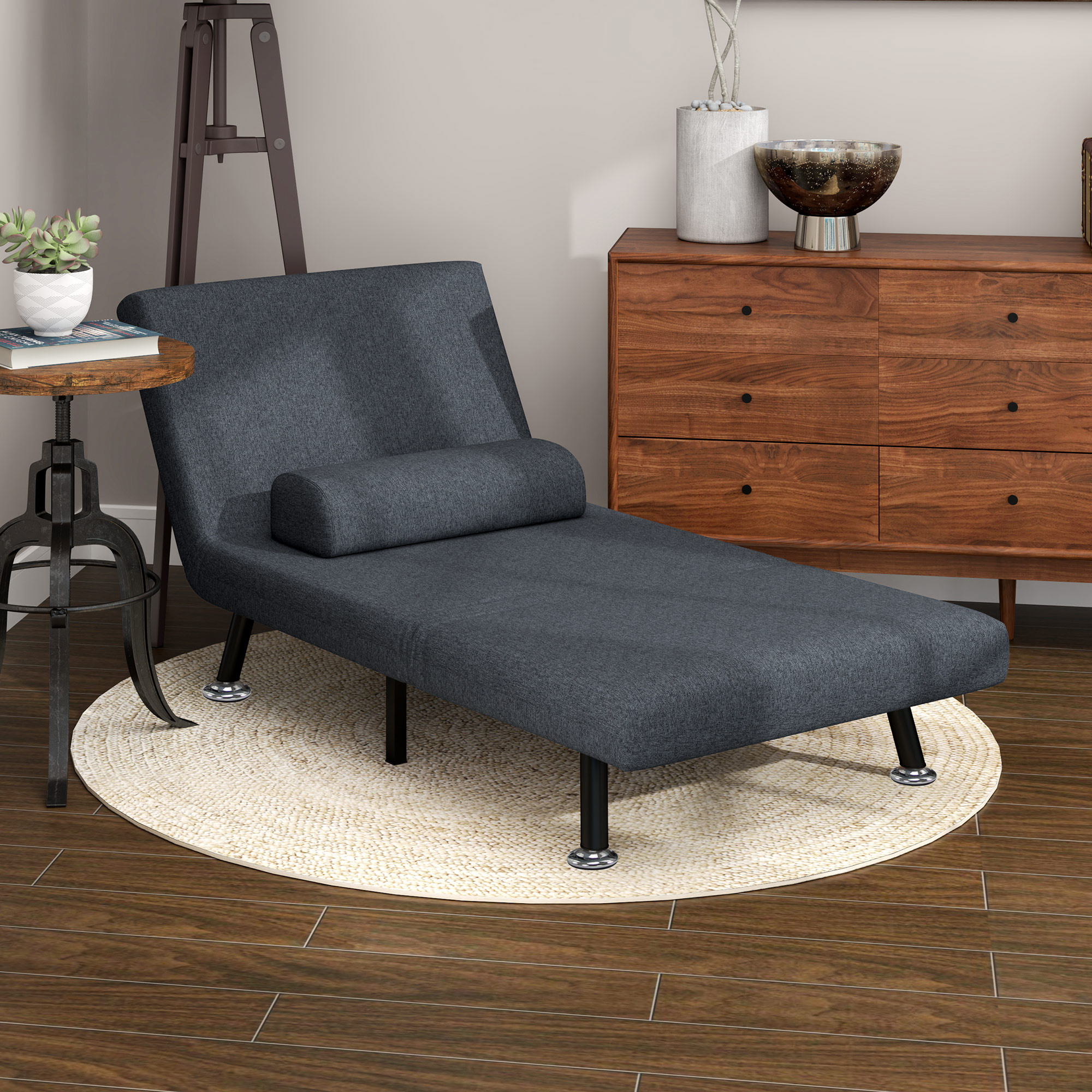 Una chaise longue grigio scuro su un tappeto tessuto. Un tavolino e una cassettiera sullo sfondo.