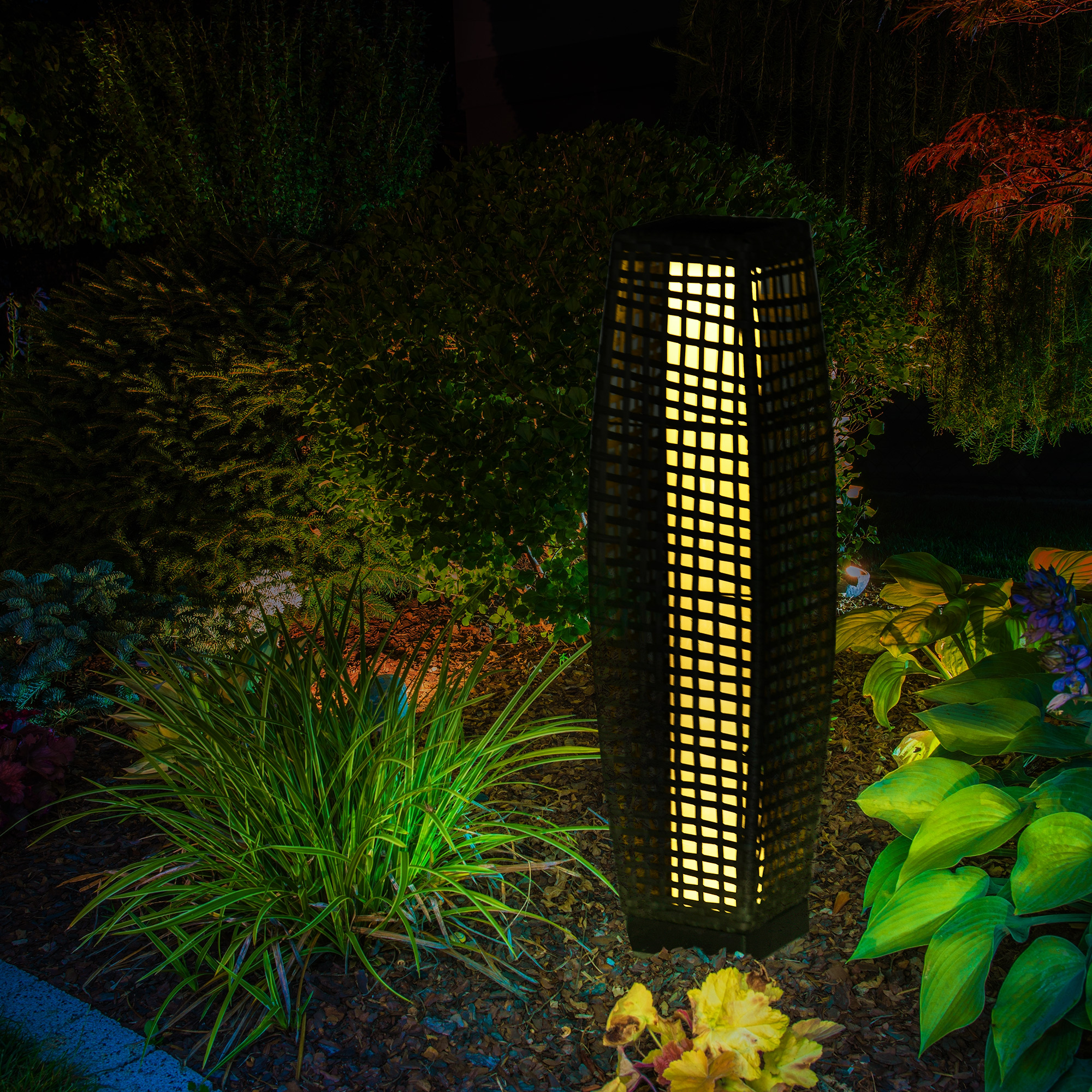 Una lampada da esterno alta e illuminata con un design a griglia, circondata da piante verdi di notte.