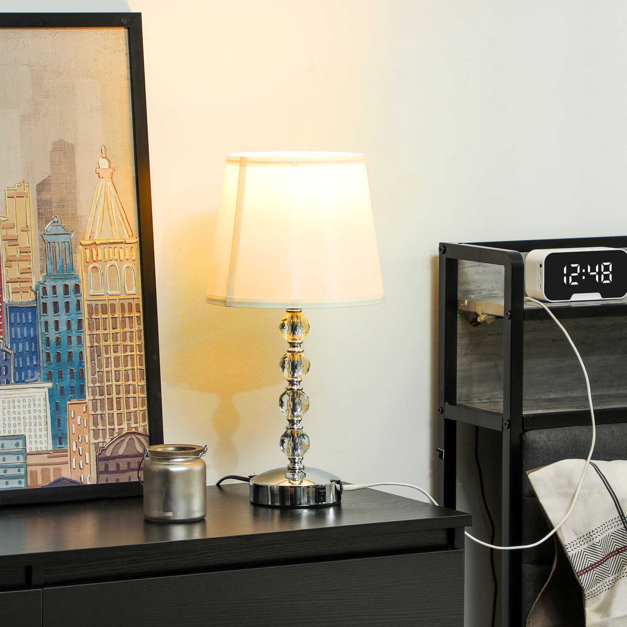 Una lampada da tavolo con paralume bianco. Un disegno di una città e un orologio digitale sono vicini.