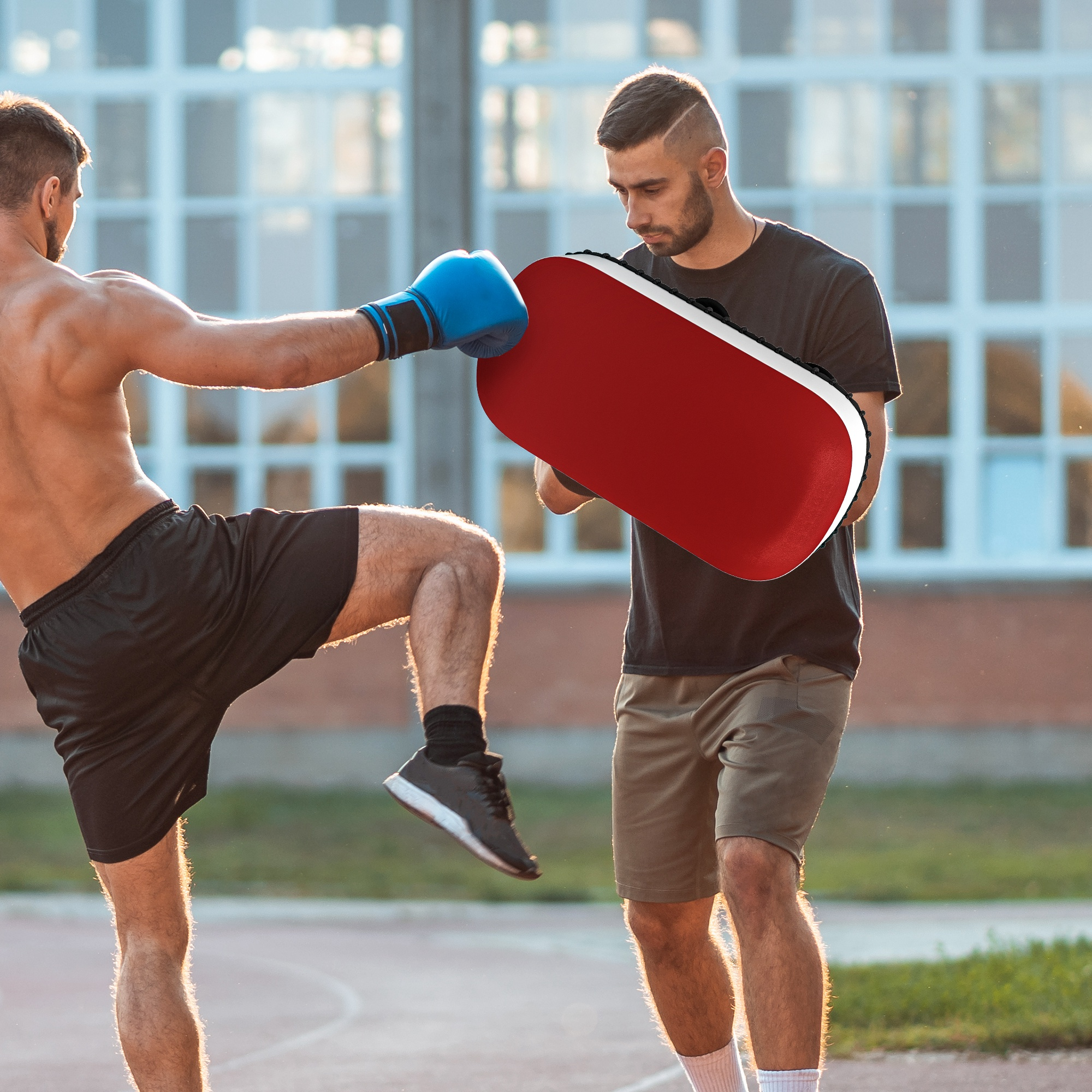 Due uomini che praticano kickboxing. Uno calcia un cuscinetto rosso tenuto dall'altro, all'aperto.