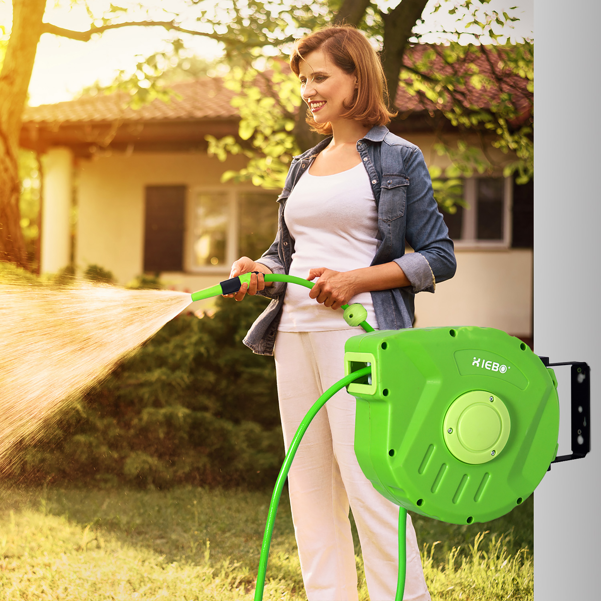 Donna che innaffia un prato con un tubo verde e un avvolgitubo verde montato a parete, giornata soleggiata.