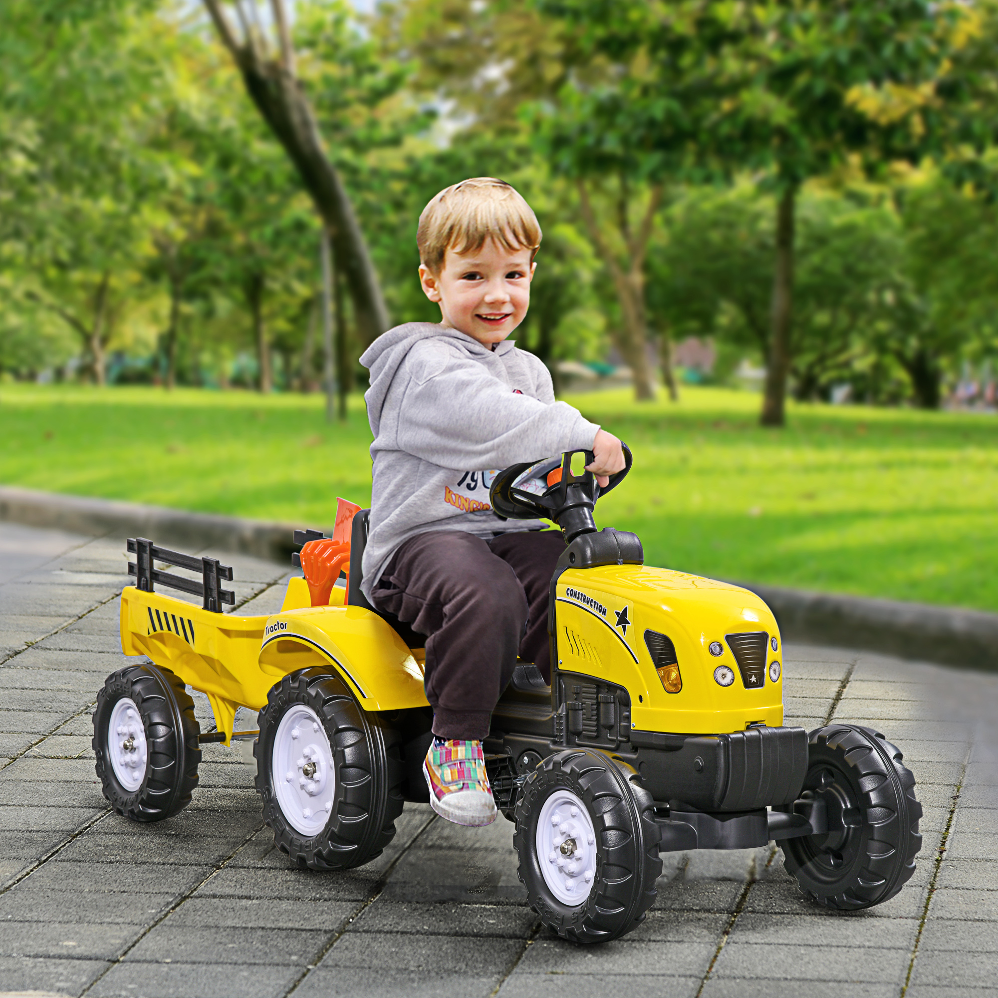 Un niño pequeño conduce un tractor amarillo por una carretera pavimentada en un parque. El tractor tiene un remolque.