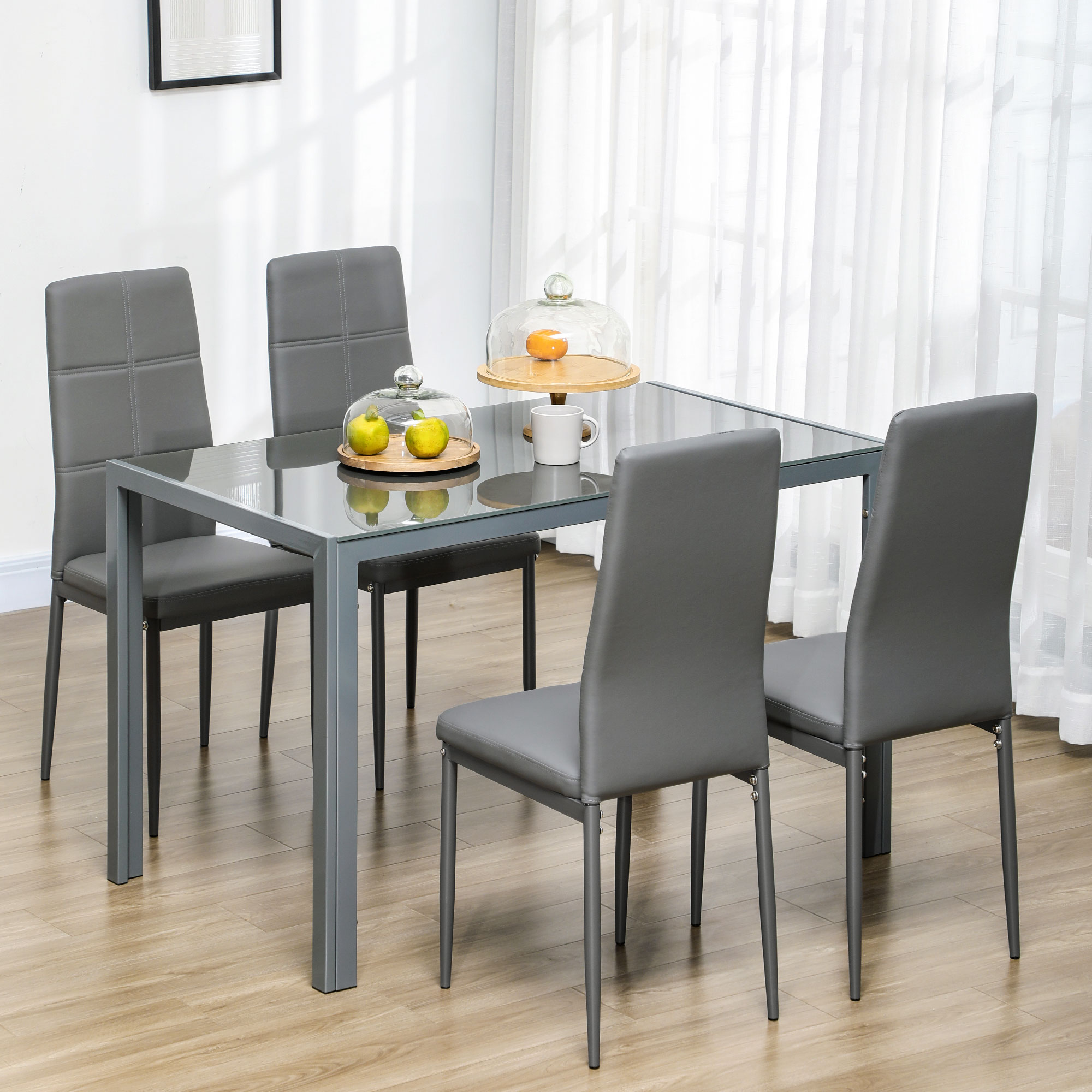 Tavolo da pranzo con piano in vetro e quattro sedie grigie. Una campana di vetro e frutta sul tavolo.
