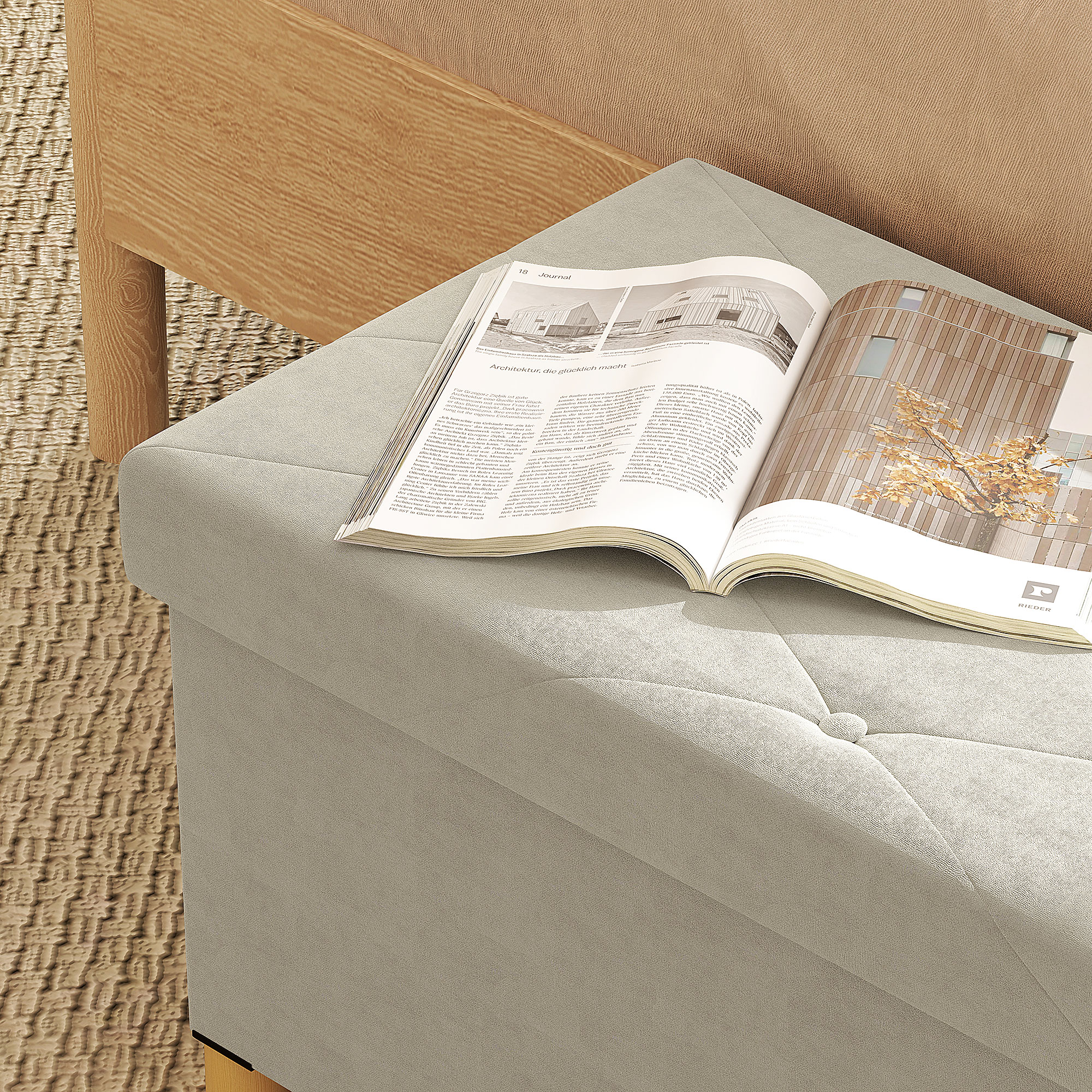 Una rivista aperta su un pouf imbottito grigio chiaro. Poggia su un tappeto fantasia e vicino a un letto.
