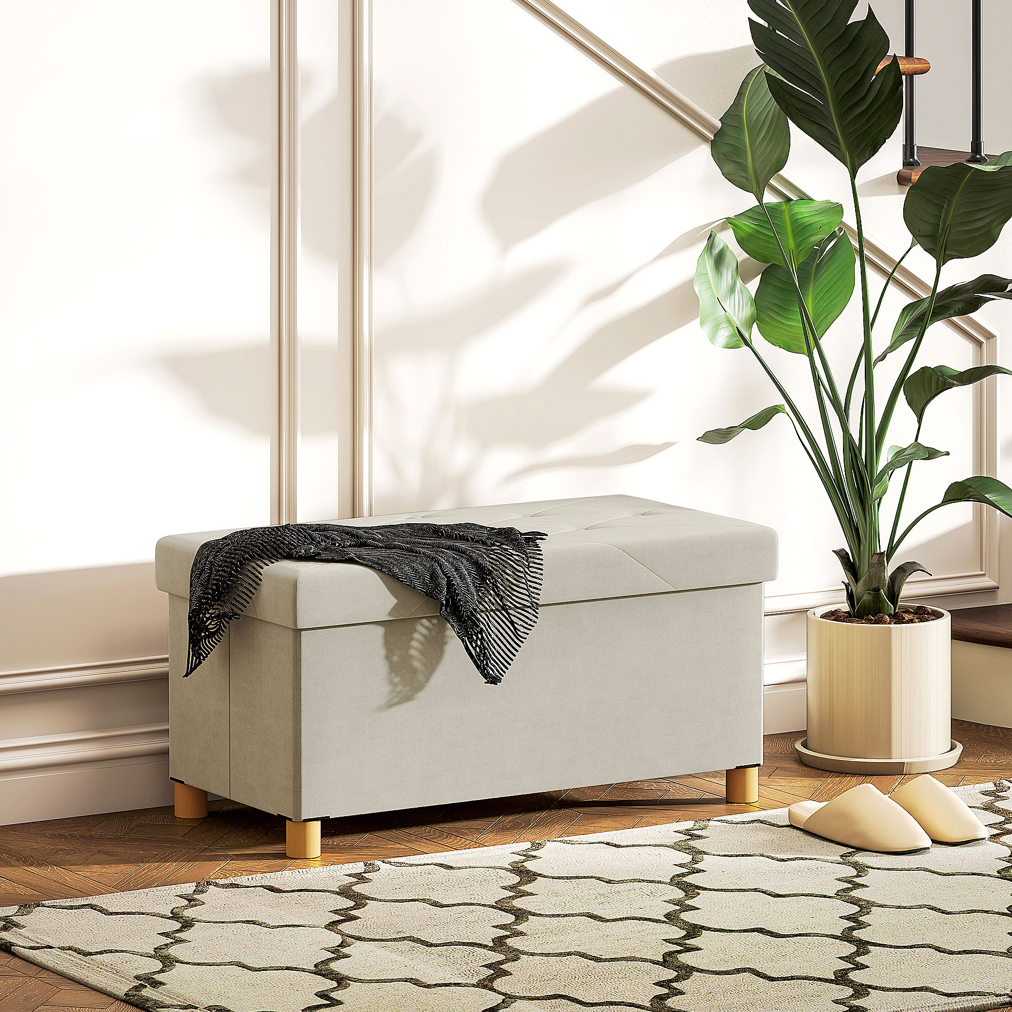 Pouf contenitore beige con coperta sopra, pianta in vaso e tappeto fantasia.