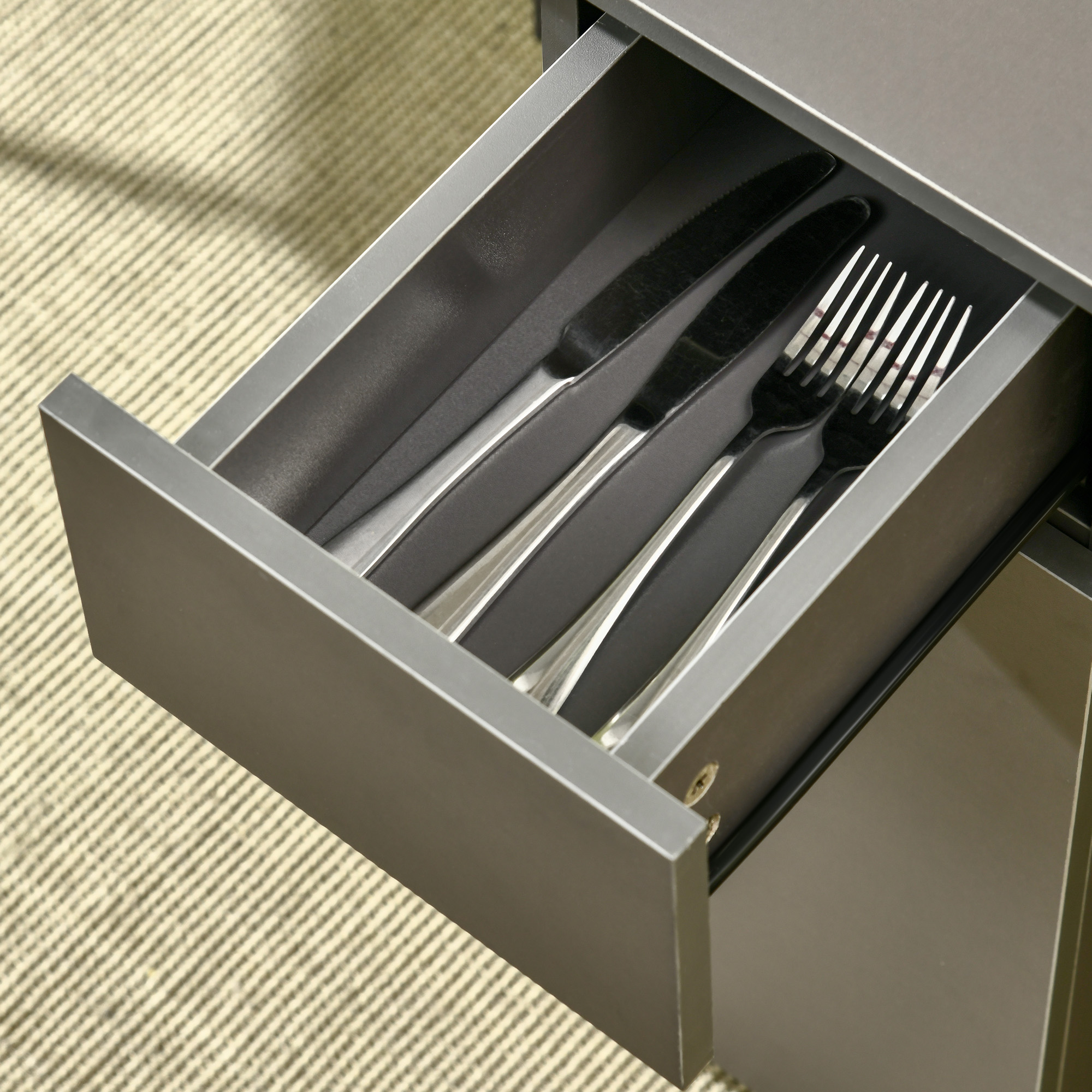 Cassetto aperto con posate: coltelli, forchette. Cassetto grigio e sfondo tappeto beige.