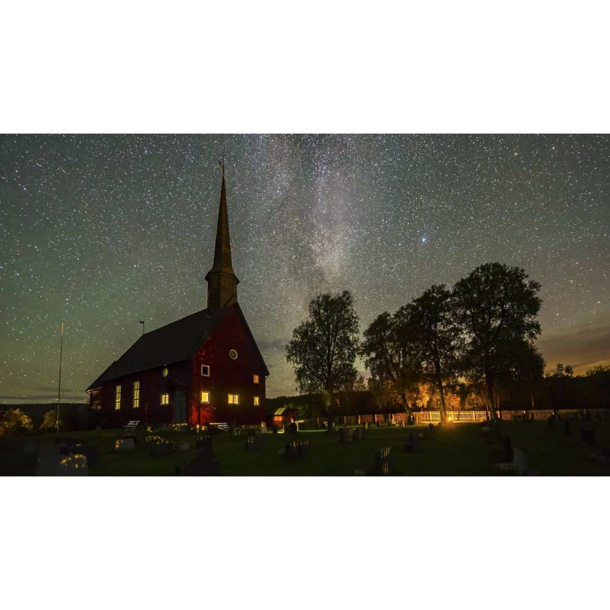 Toma nocturna de una iglesia con ventanas brillantes y cielo estrellado. Árboles y un cementerio a la vista.
