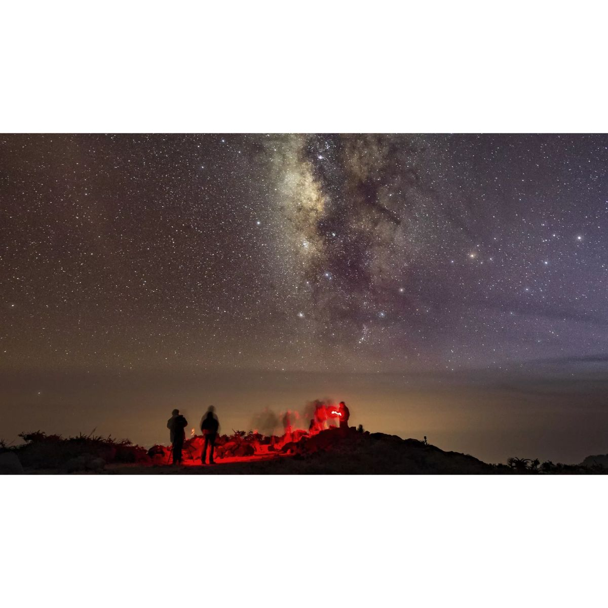 Escena nocturna con personas alrededor de luz roja, bajo cielo estrellado.