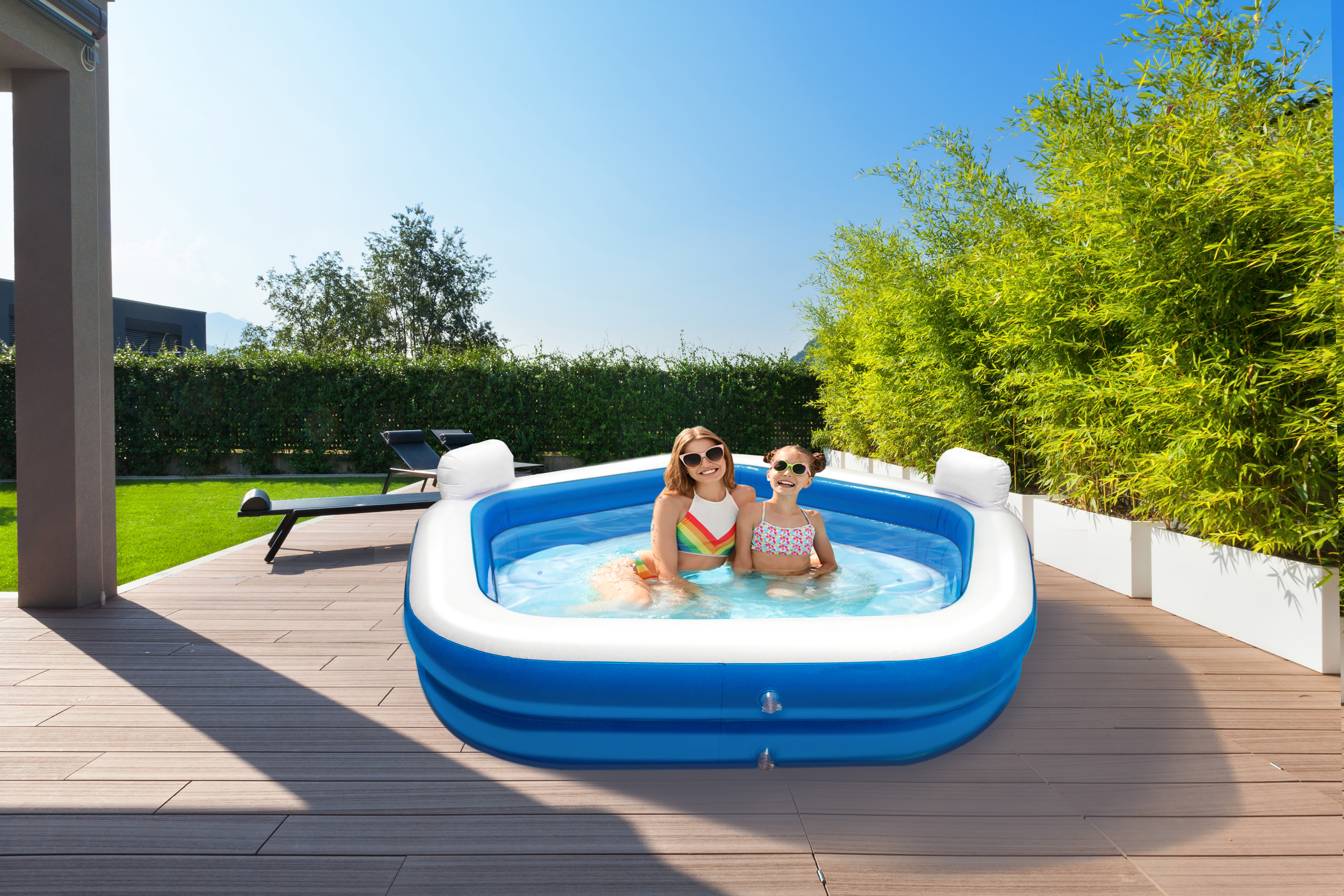 Twee meisjes in badpakken zitten in een blauw-wit opblaasbaar zwembad op een houten terras, zonnige dag.