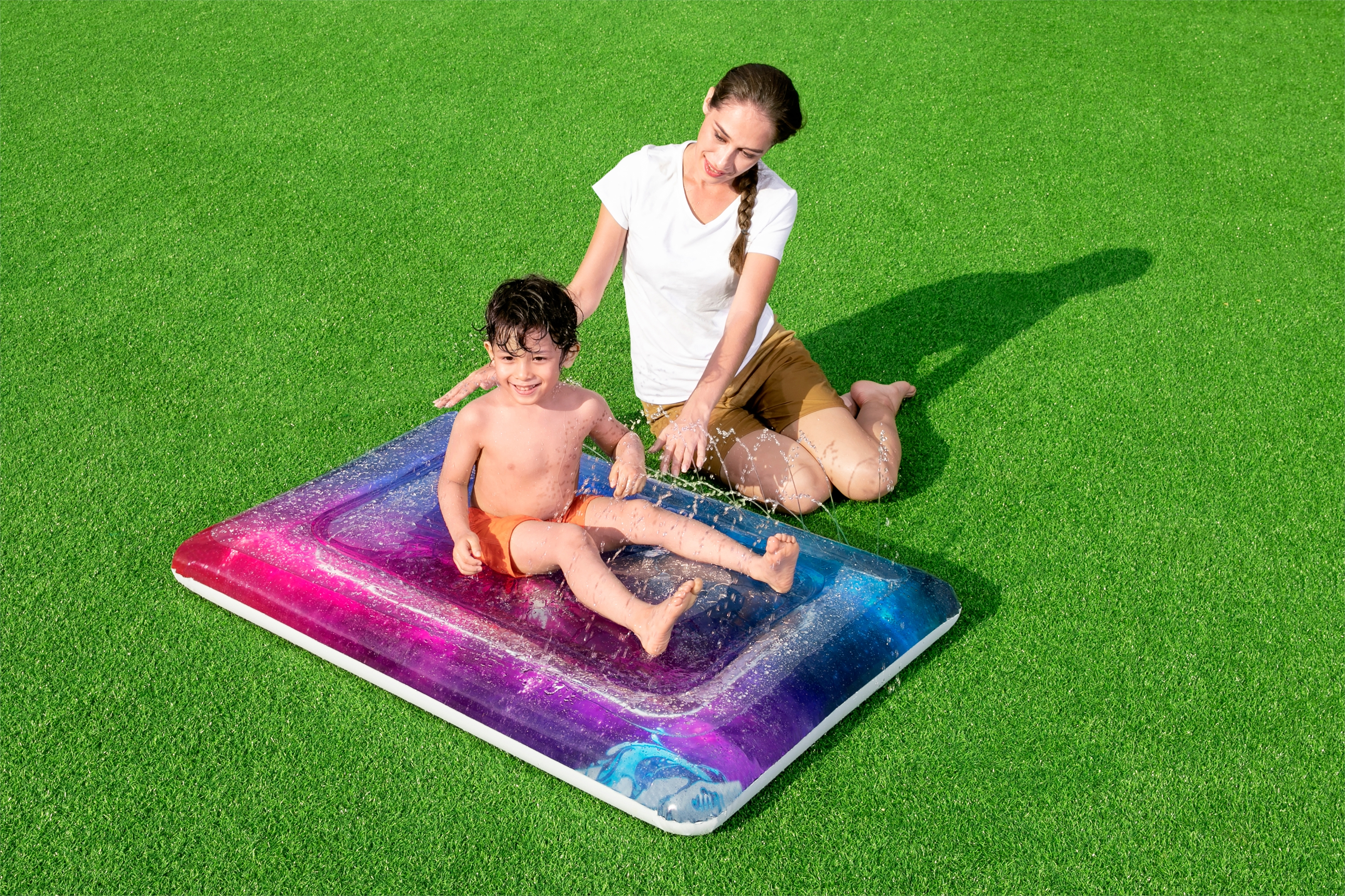 Een kind en een vrouw die op een sproeimat spelen. De mat is kleurrijk en ligt op groen gras.