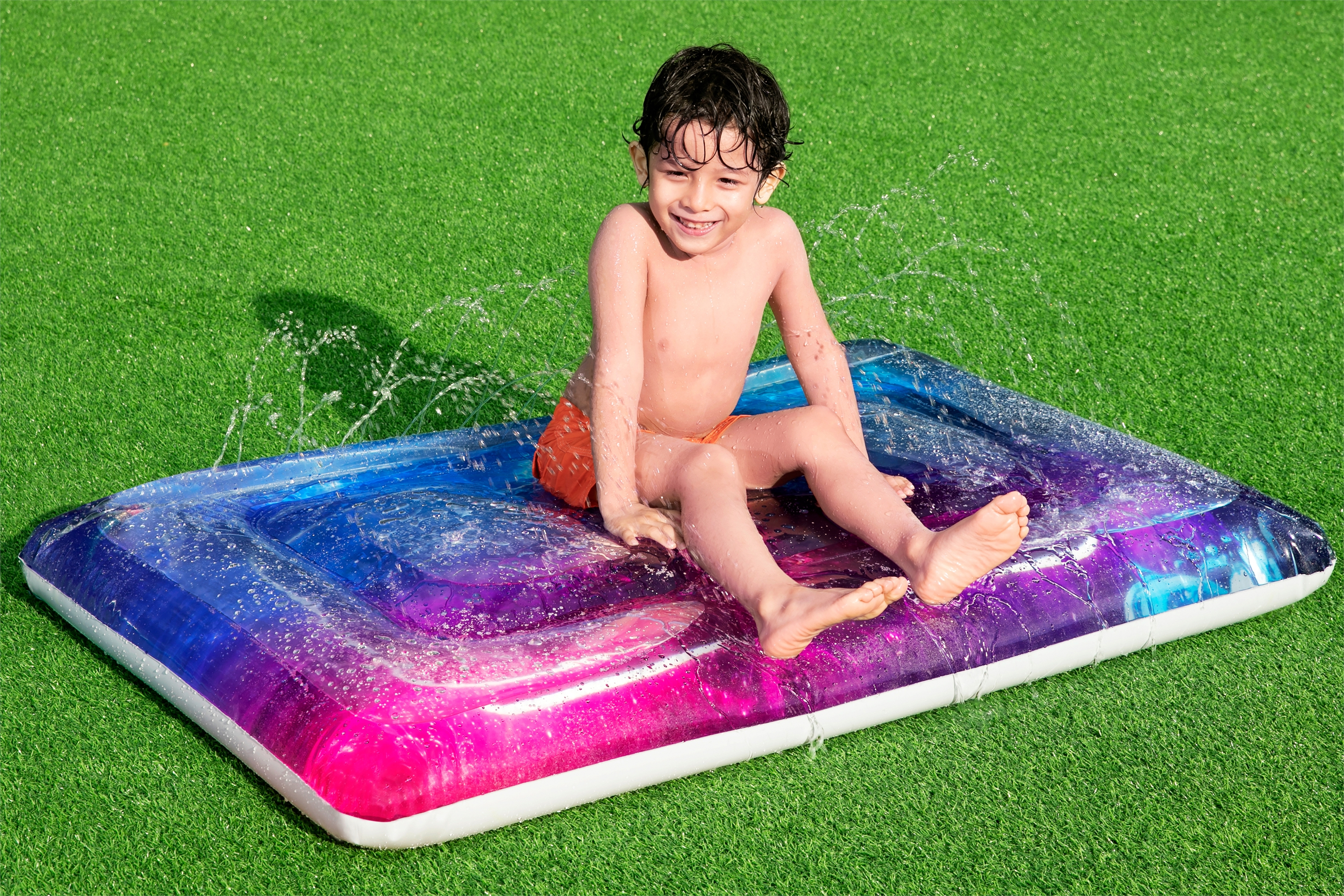 Een jongen op een kleurrijke opblaasbare matras op groen gras, met opspattend water.