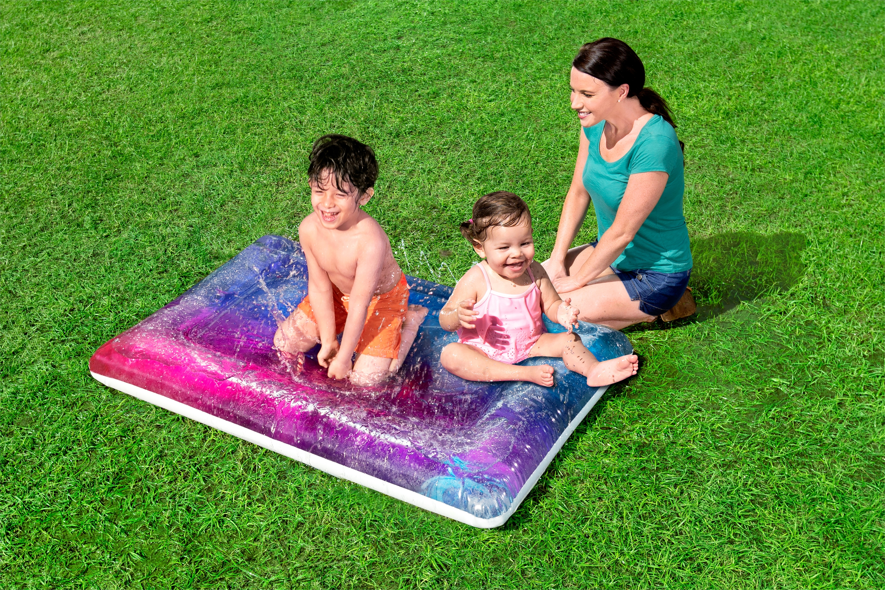 Drie mensen spelen op een watermat op het gras. Een volwassene en twee kinderen glimlachen.