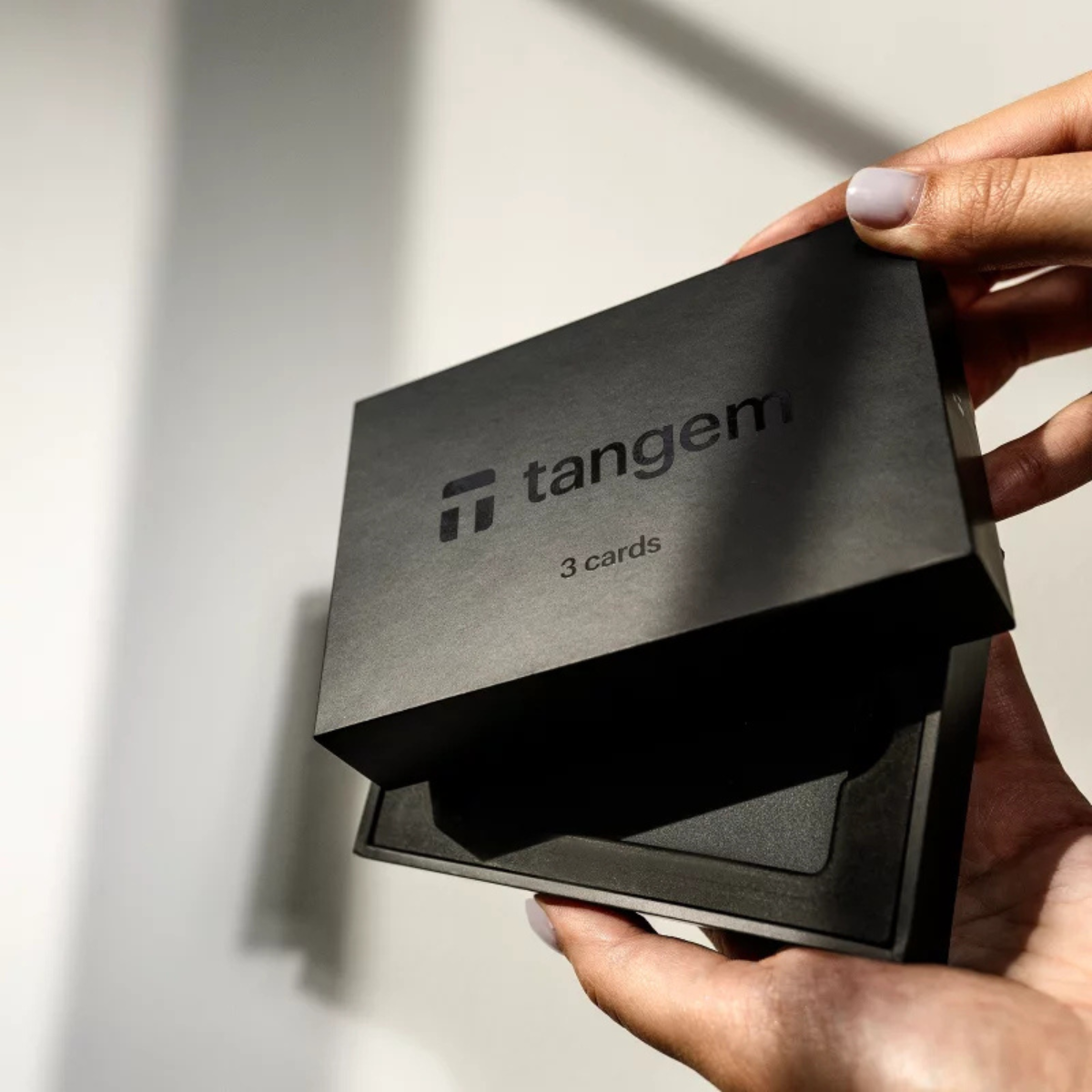 Dłonie otwierające czarne pudełko z logo Tangem, ukazujące karty w środku, białe tło.
