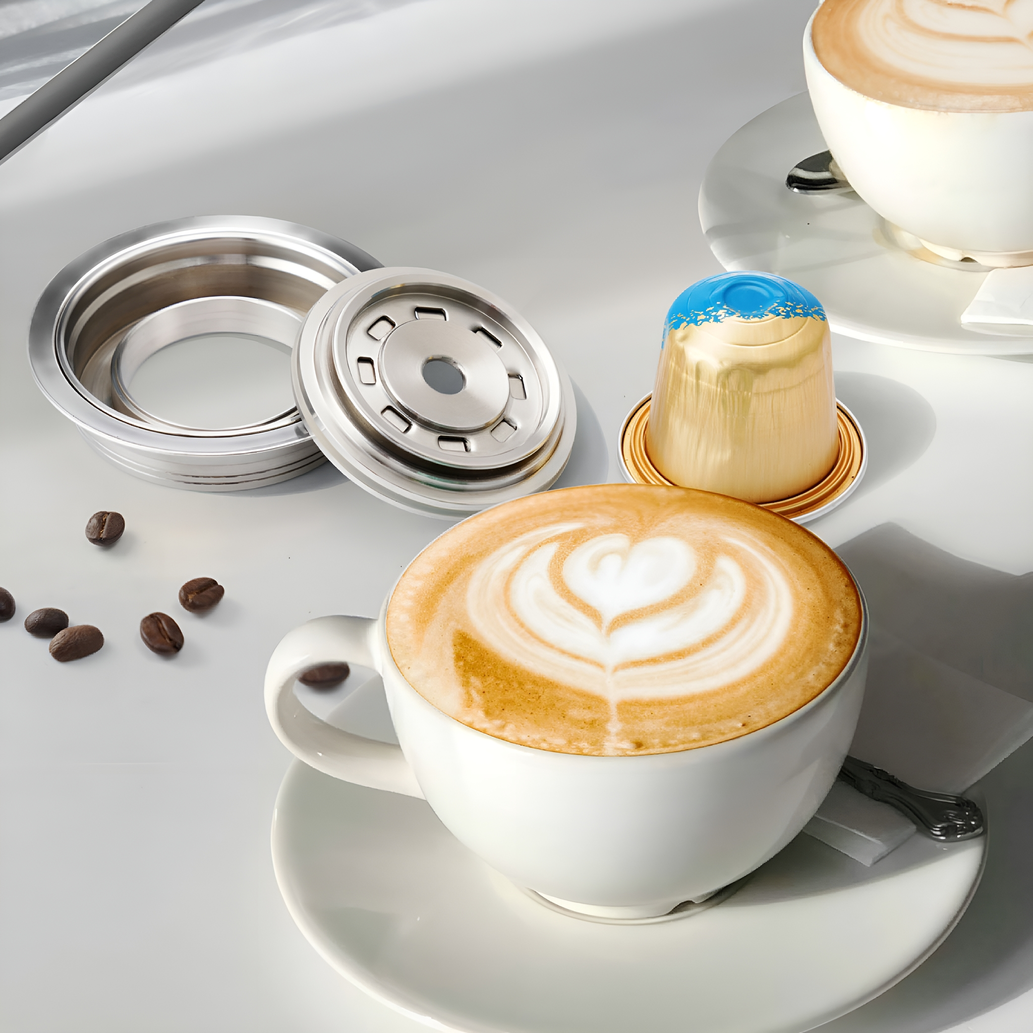 Zwei Tassen Latte Macchiato mit herzförmiger Schaumkunst, eine Kaffeekapsel und Kaffeebohnen auf weißer Oberfläche.