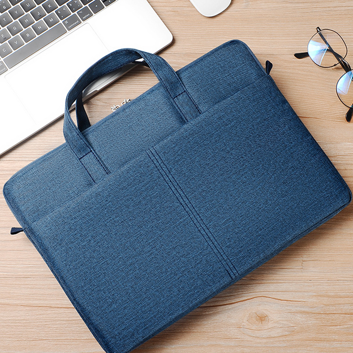 Eine blaue Laptoptasche mit Griffen, liegt auf einem Holztisch neben einem Laptop und einer Brille.