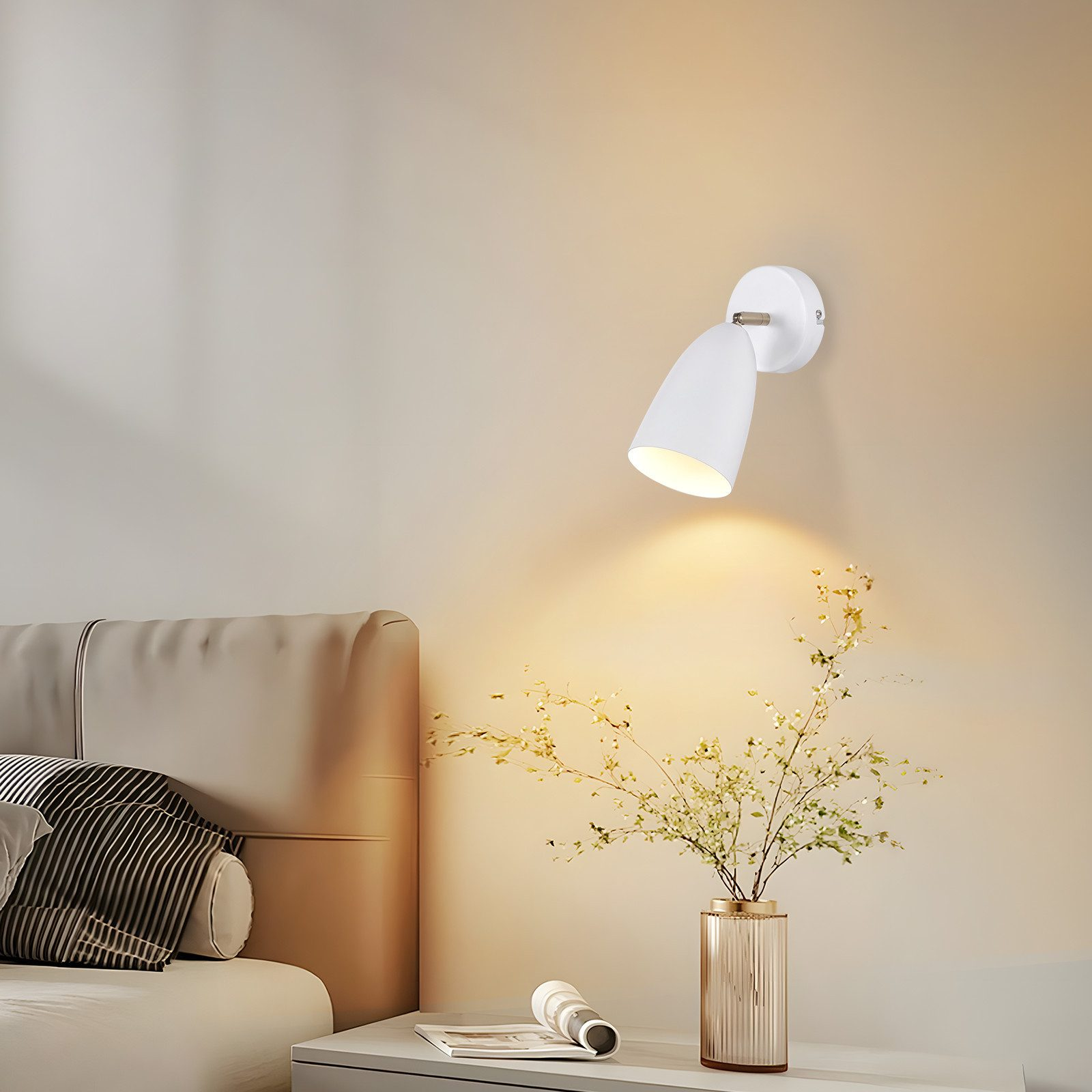 Una lampada bianca a parete con un design a faretto. Un vaso di fiori è posto sotto su un tavolo.