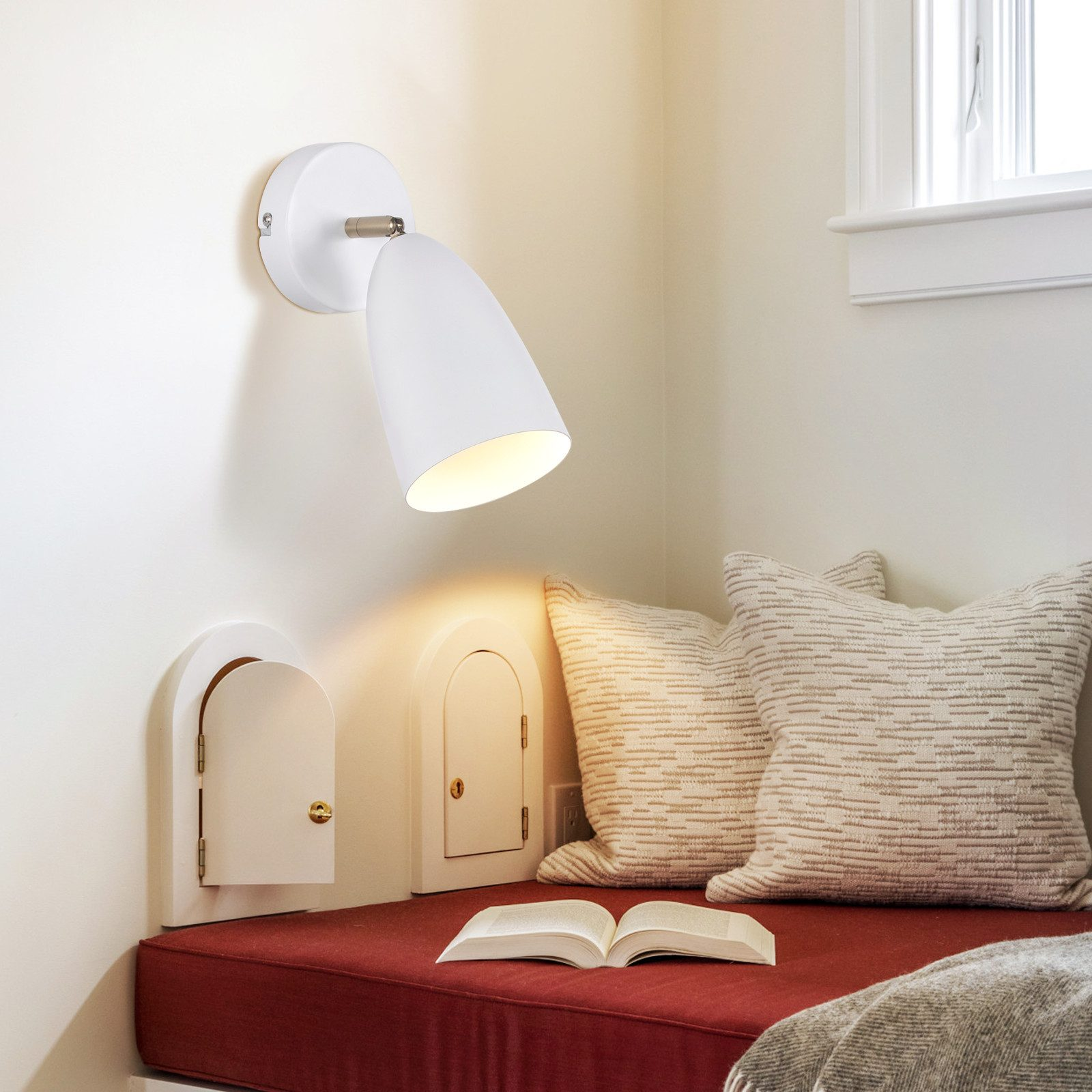 Lampada bianca a parete illumina cuscino rosso e libro aperto.