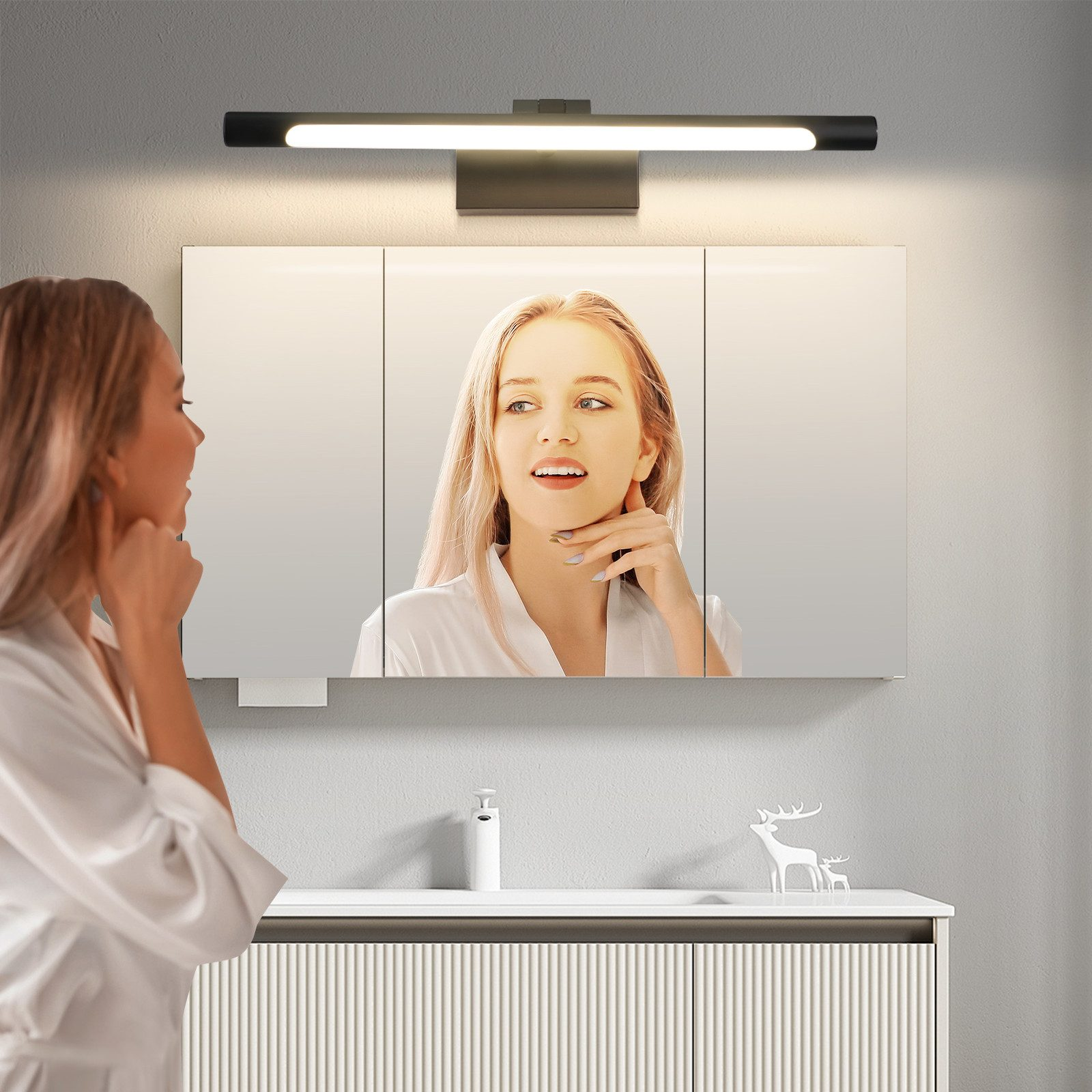 Una donna in accappatoio bianco si guarda in uno specchio, luce del bagno sopra.