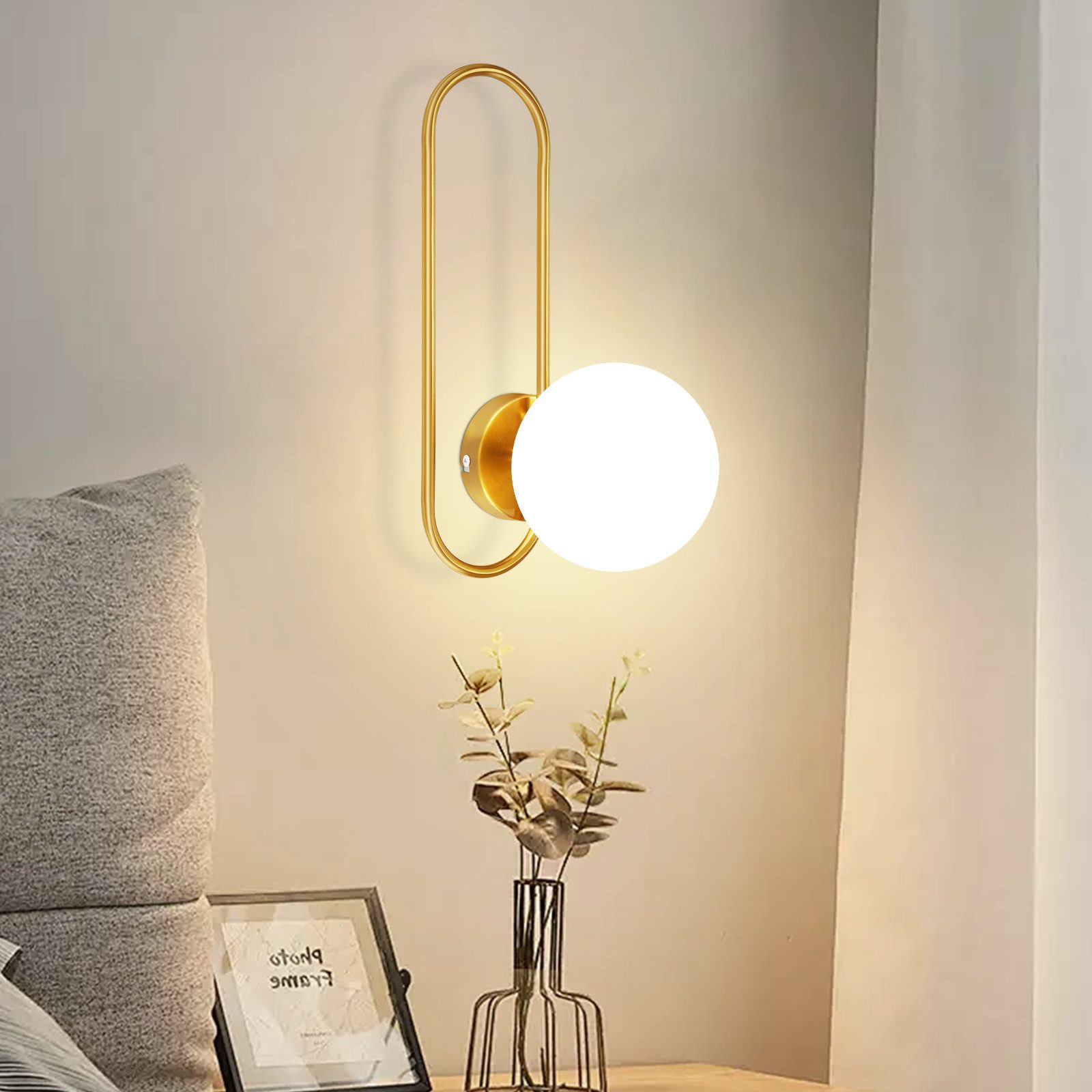 Una lampada da parete con una cornice dorata e un globo bianco. Un vaso di fiori e una cornice per foto sotto.