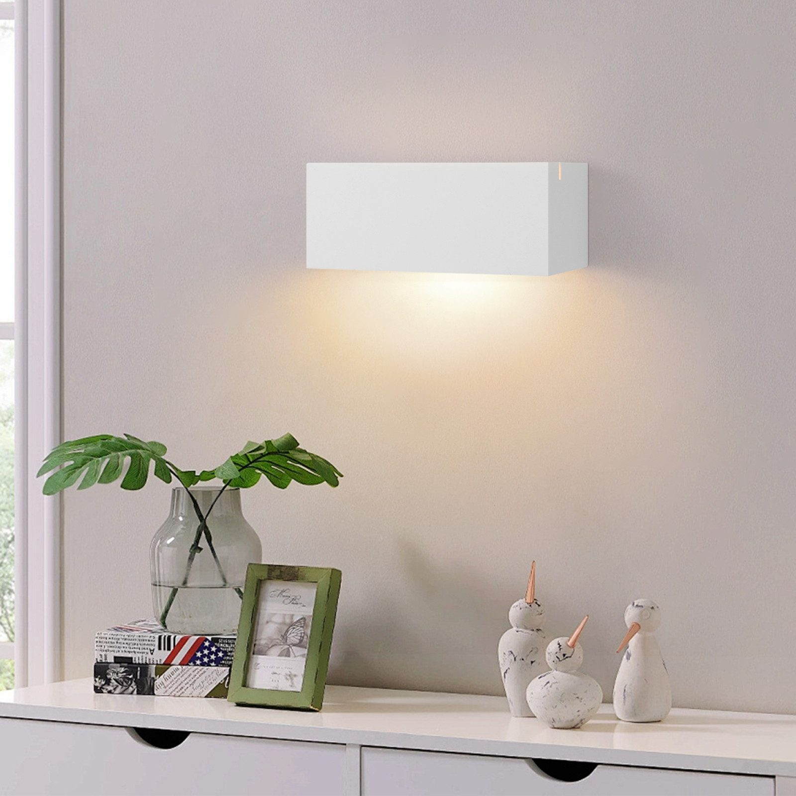Lampada da parete rettangolare bianca sopra un mobile bianco con piante e decorazioni.