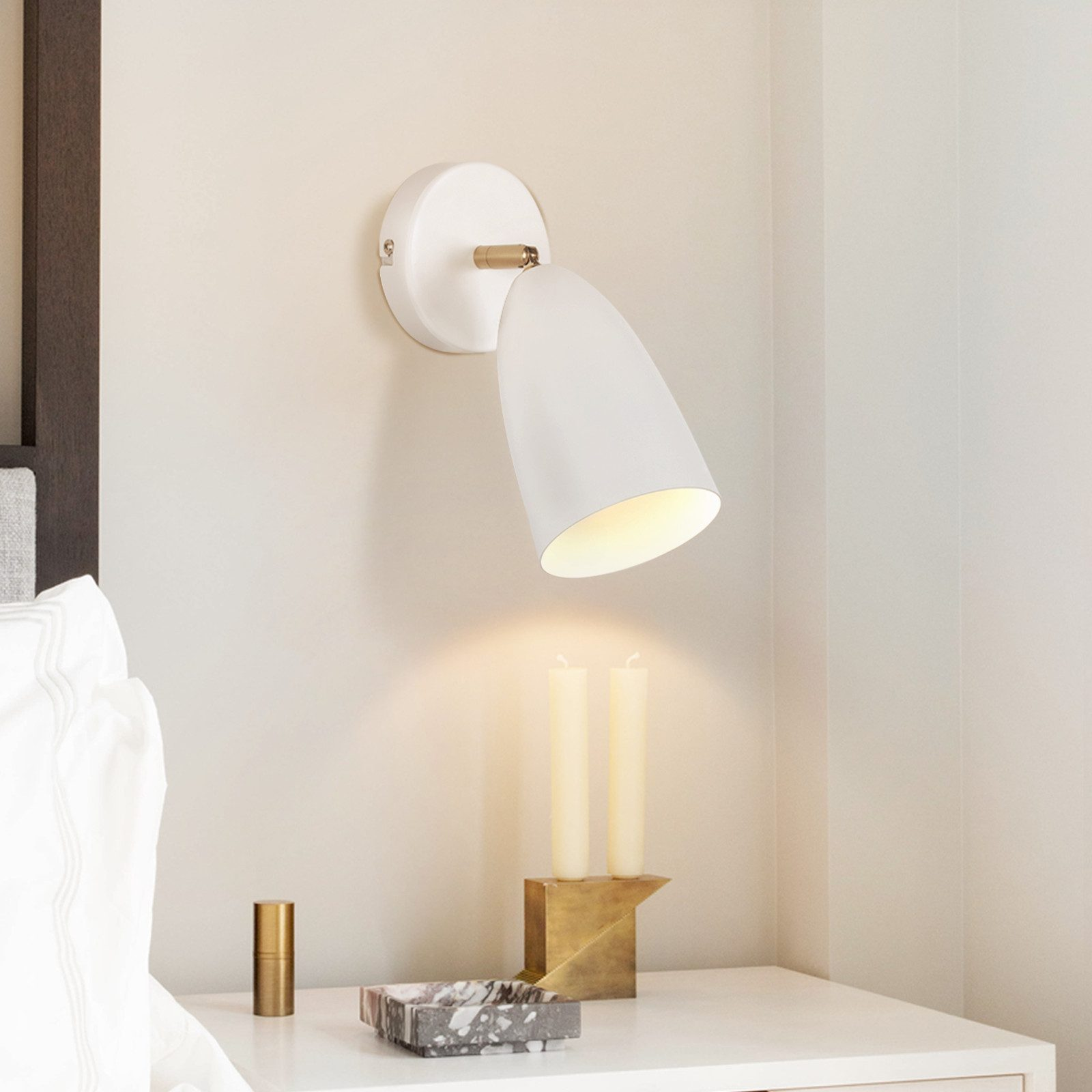 Una lampada da parete bianca con un faretto e due candele su un tavolo accanto a un muro beige.