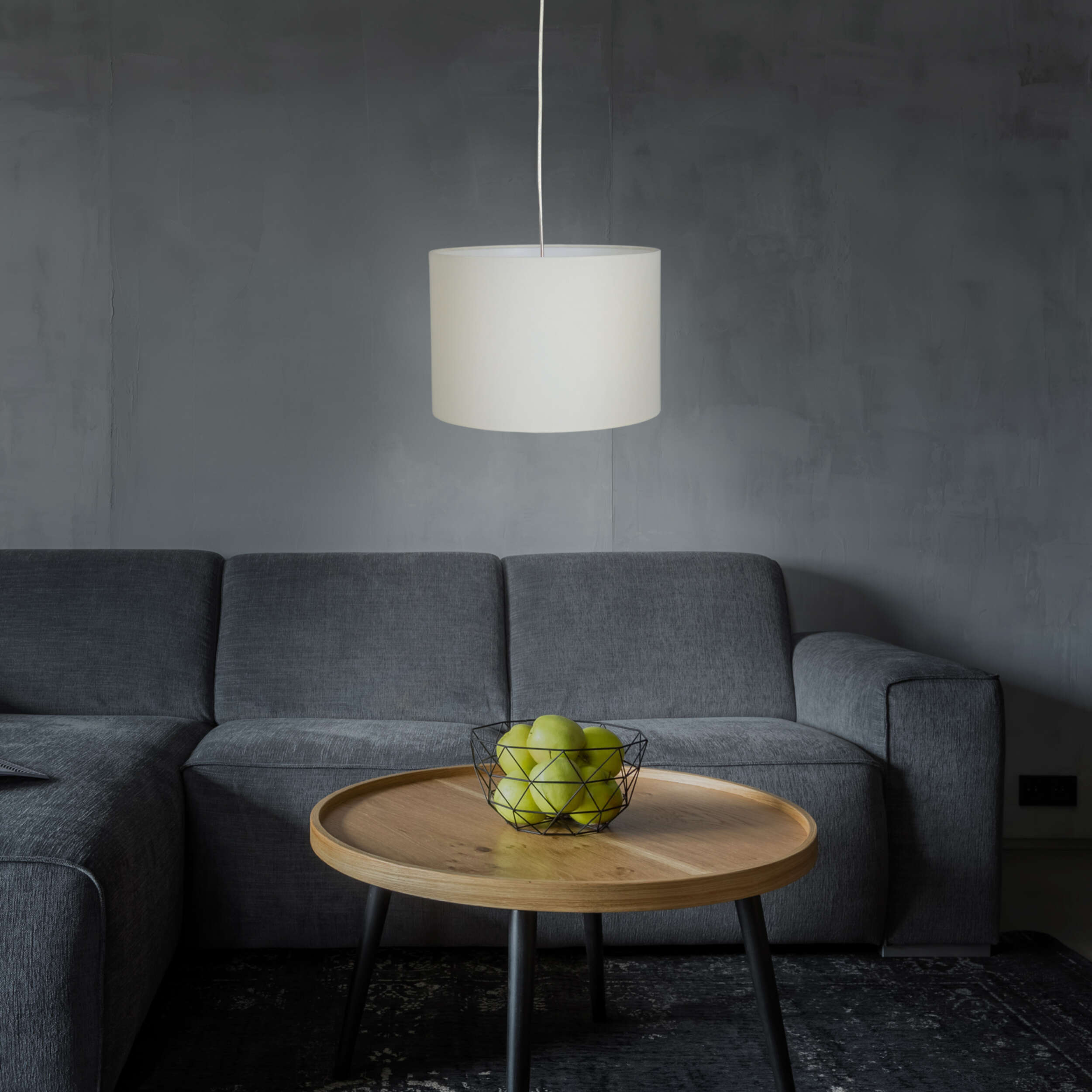 Eine weiße zylindrische Lampe hängt über einem Holztisch mit einer Schale grüner Äpfel, graues Sofa.