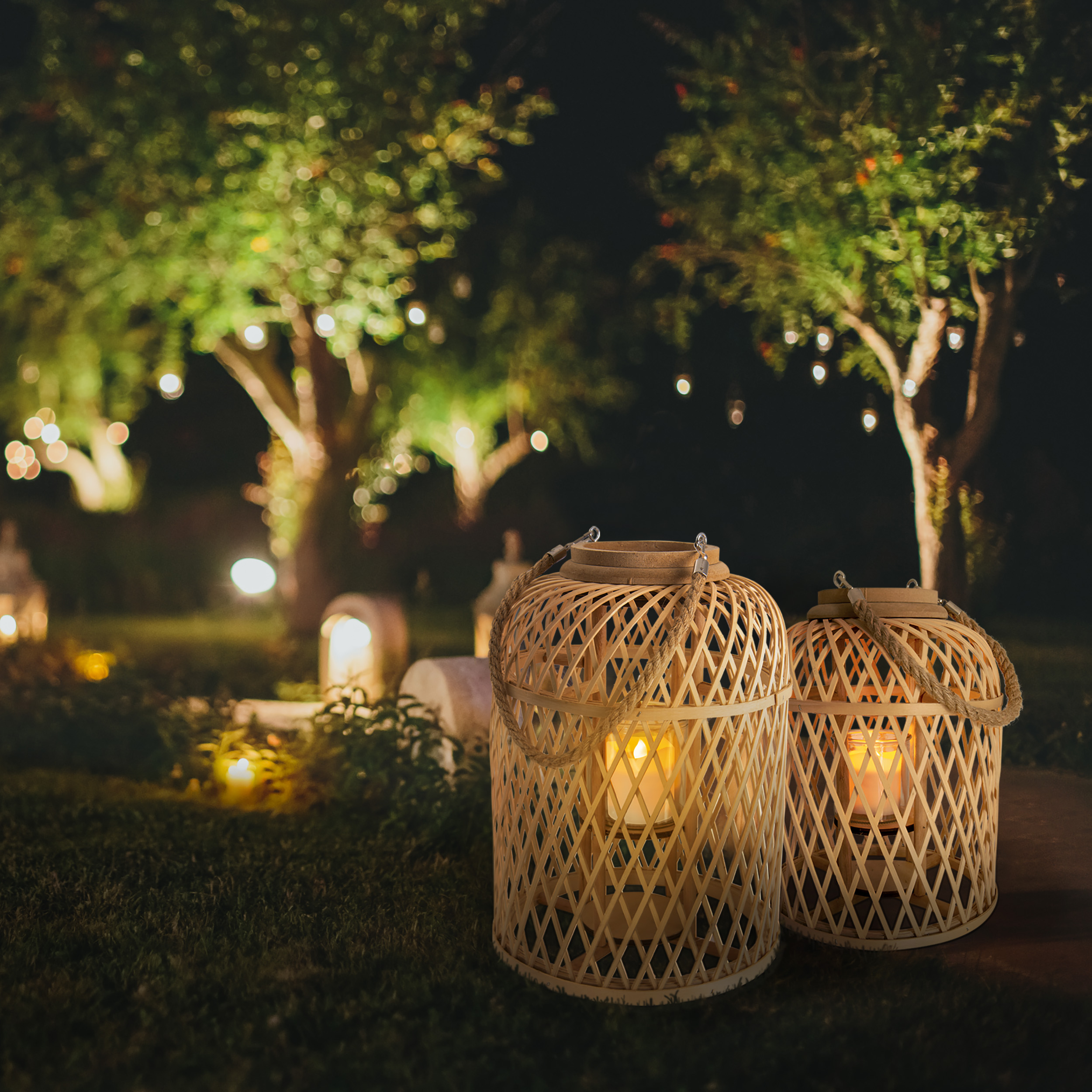 Zwei beleuchtete Bambuslaternen mit Kerzen in einem Garten bei Nacht mit Bäumen und Lichterketten im Hintergrund.