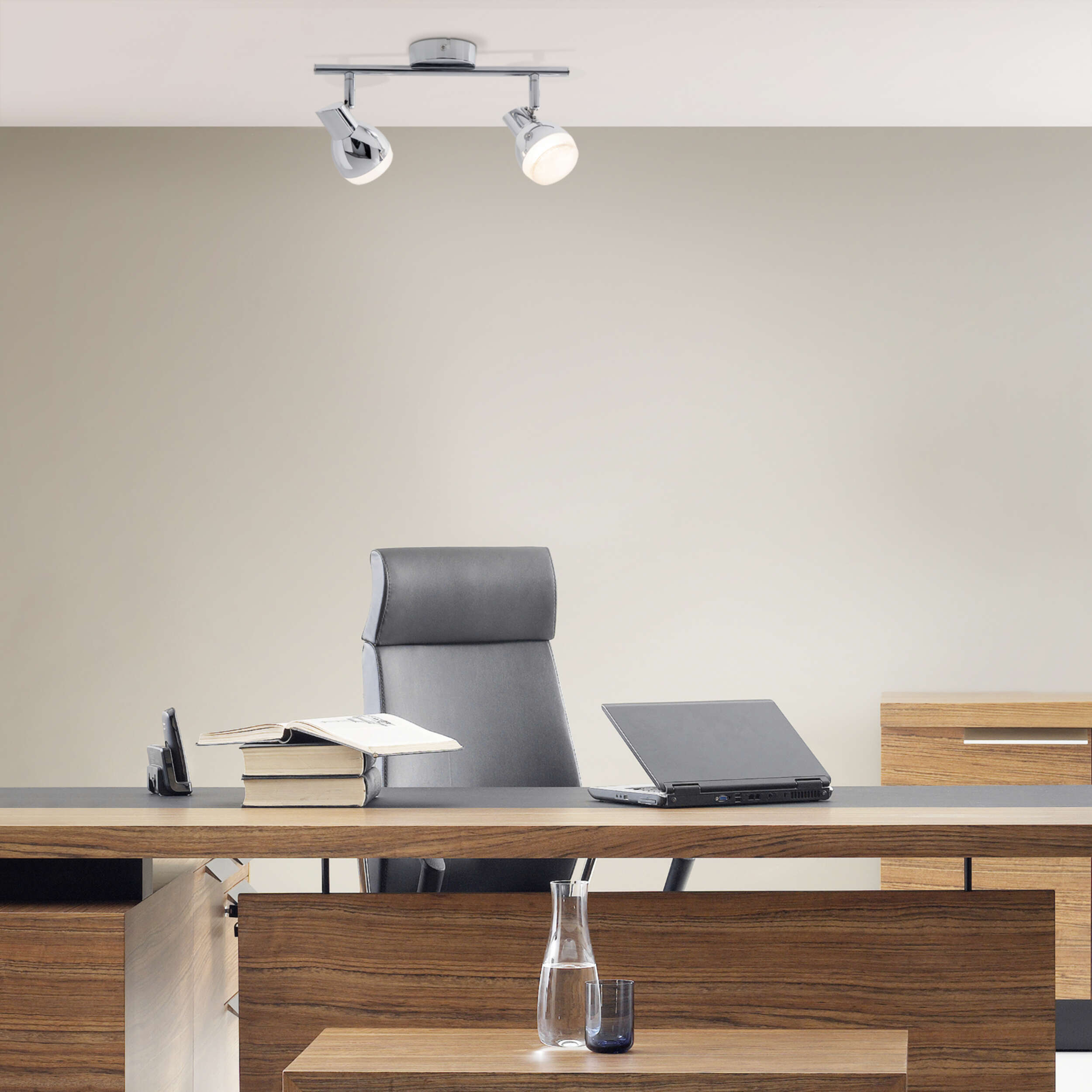 Bürotisch mit Laptop, Büchern und Wasserflasche. Deckenstrahler beleuchten die Szene.