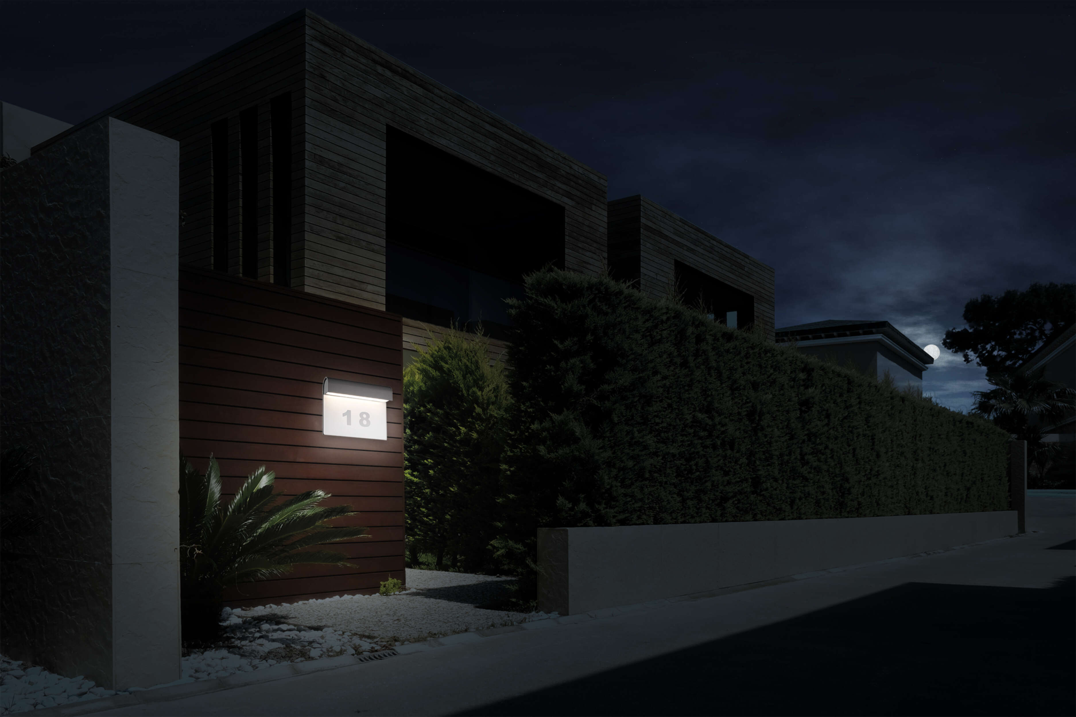 Hausfassade bei Nacht mit beleuchteter Hausnummer 18 an der Wand und hellem Licht.